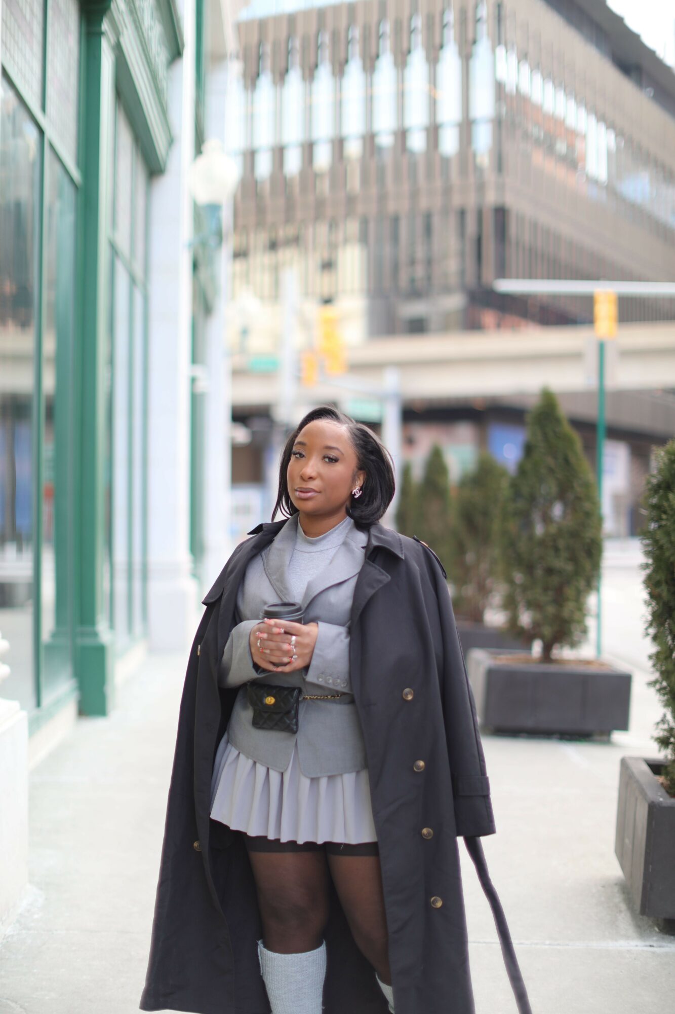 Woman standing outdoors in a city, wearing a coat, skirt, and holding a bag, with buildings and trees in background.