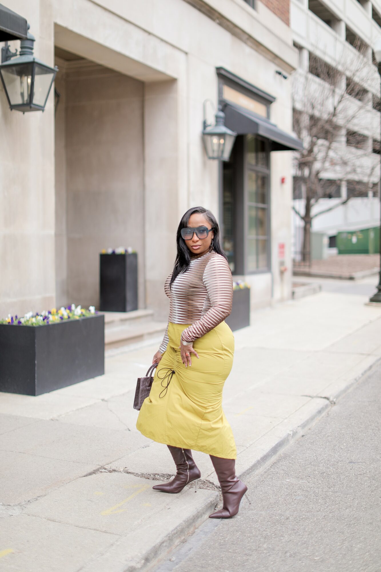Woman standing on sidewalk in front of building, wearing sunglasses, striped top, yellow skirt, and brown boots, holding a bag.
