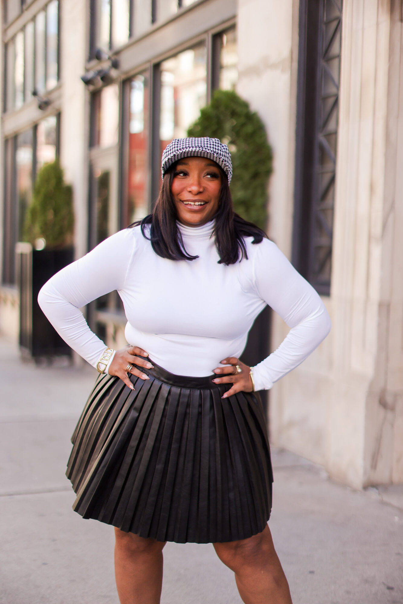 Woman standing outdoors in front of buildings, wearing a white long-sleeve top, black pleated skirt, and a patterned hat, smiling.