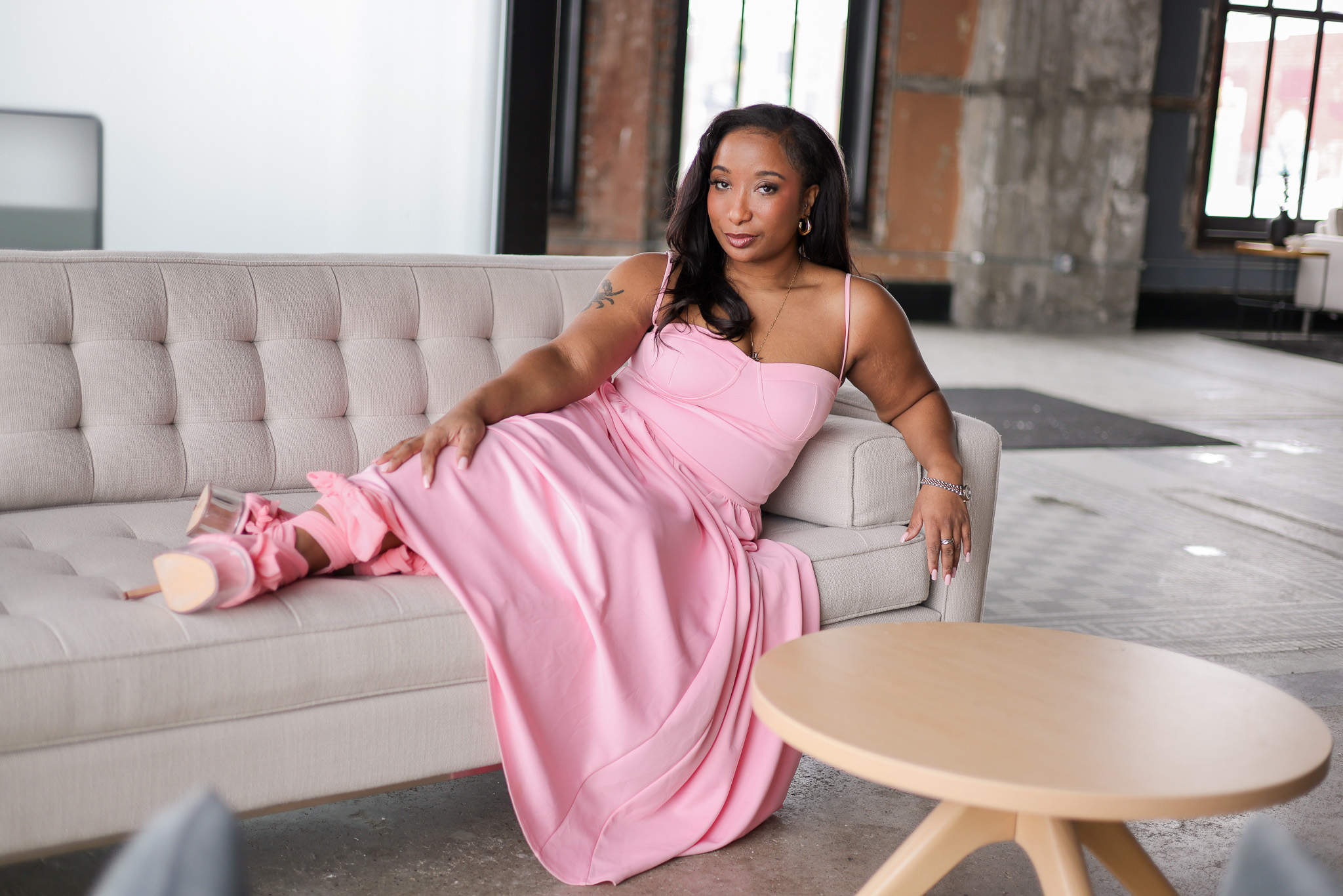 Woman in pink dress reclining on a light-colored sofa in an industrial-style room with large windows.