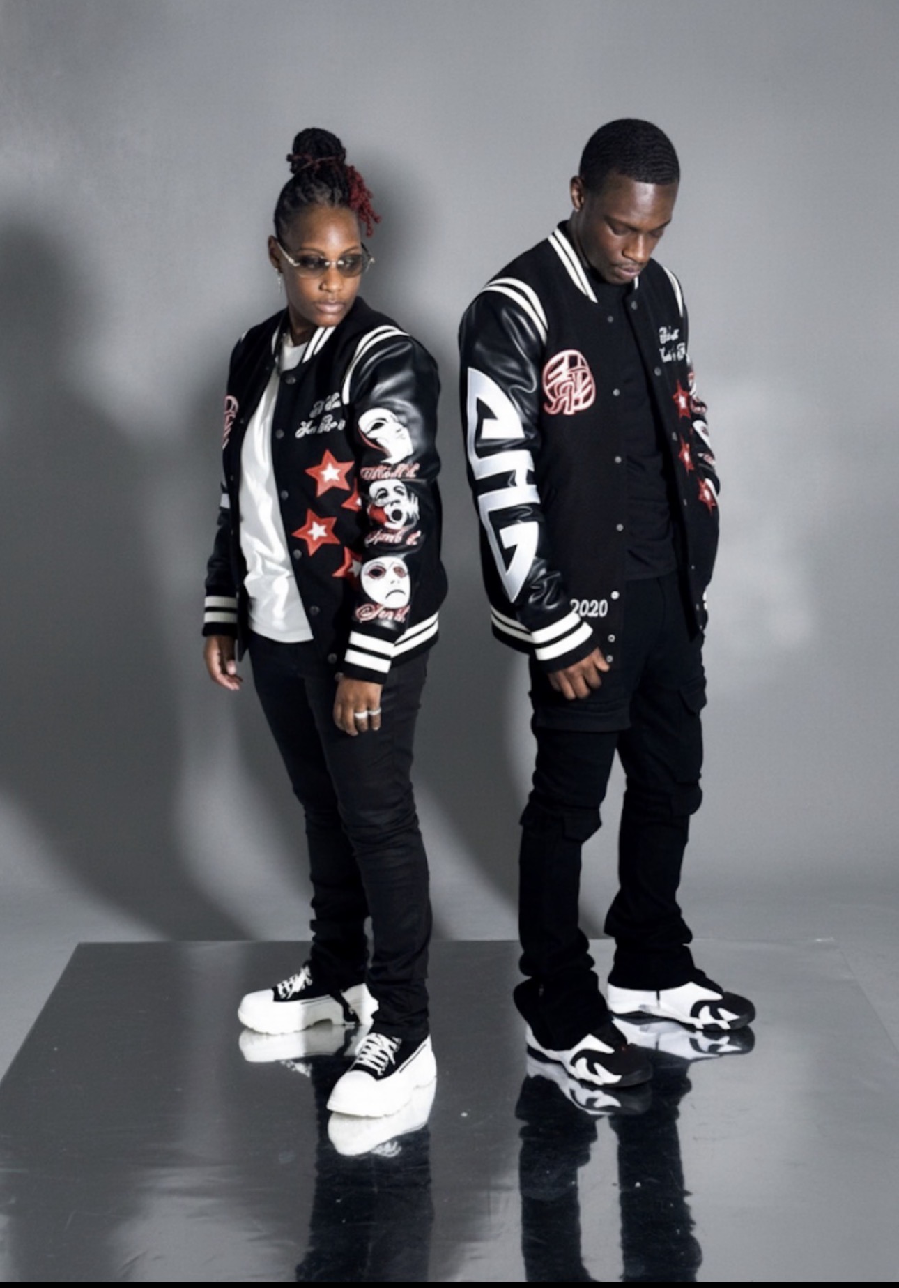 Two young people in black and white varsity jackets with patches, standing on a reflective surface against a gray background.