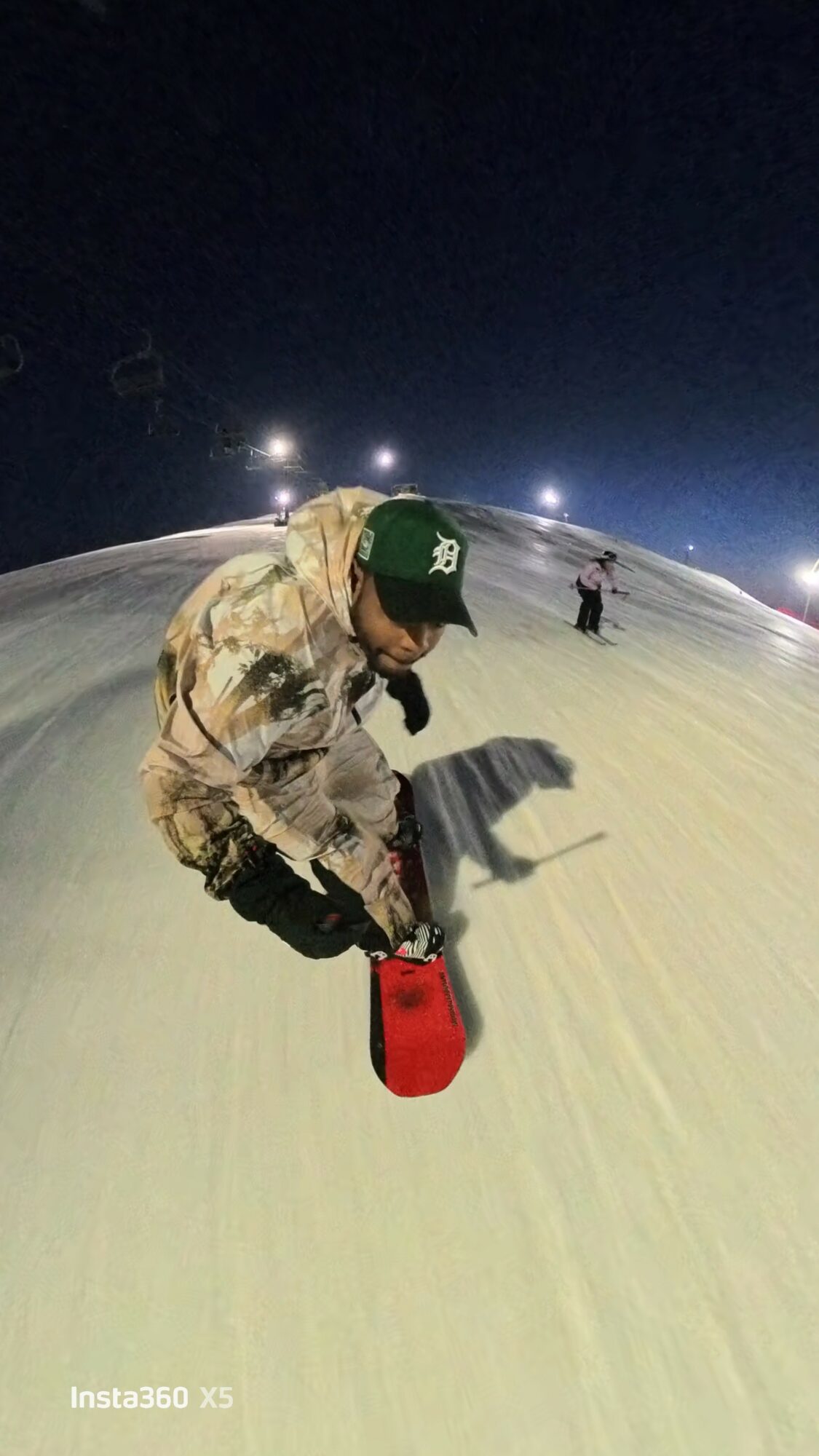 Person in camouflage jacket and green cap kneels on snow, holding a red container, with other people and lights in background.