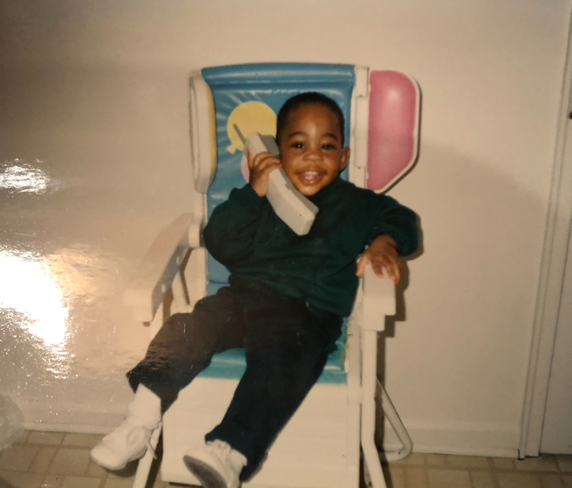 Child sitting on a colorful chair, smiling, holding a phone to ear, wearing dark clothing and white shoes.