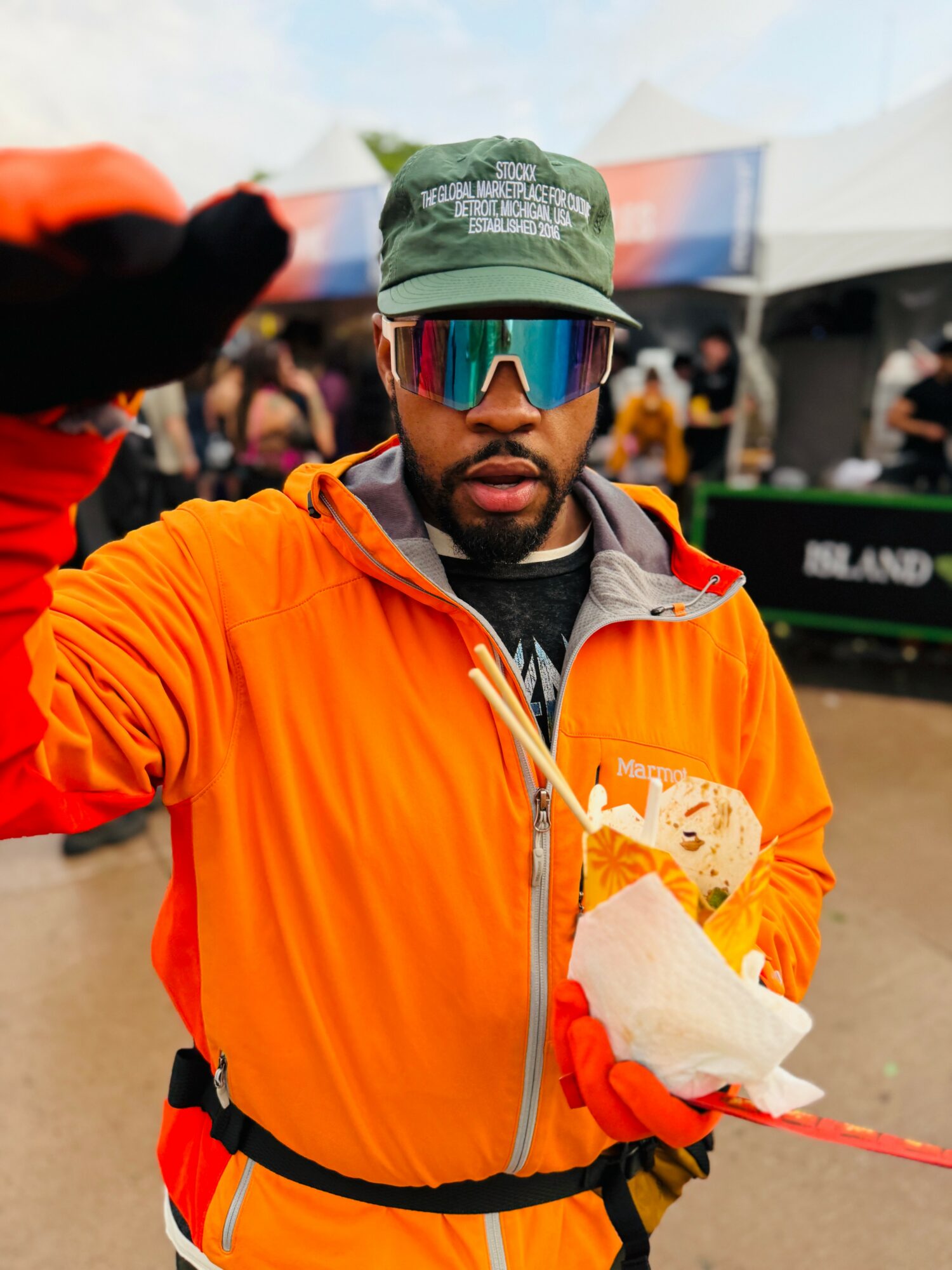 Man wearing sunglasses and a green cap, holding food, outdoors with tents and people in background.