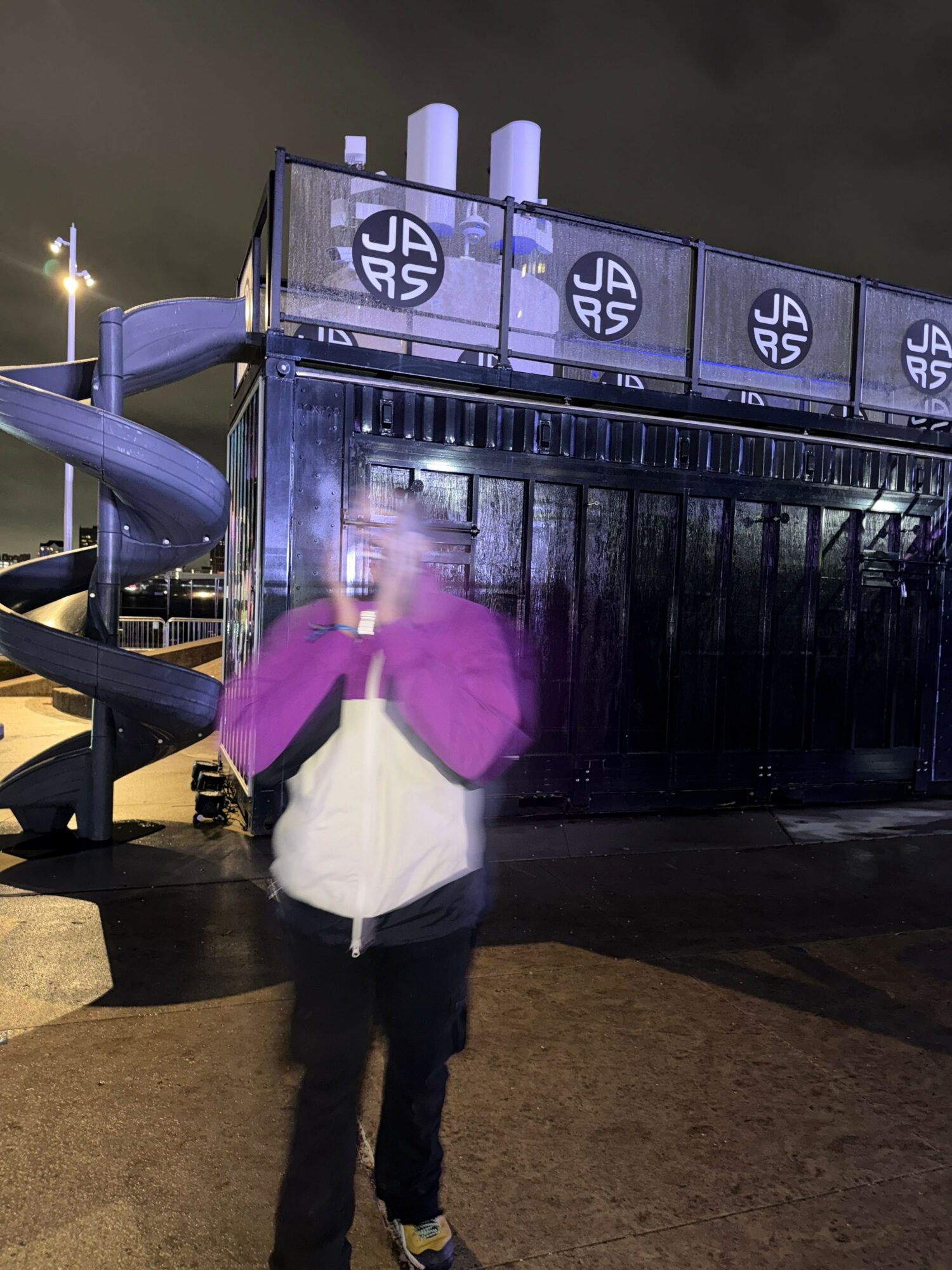 Person wearing a purple jacket and white shirt standing near a black metal structure with white symbols, in an indoor setting.