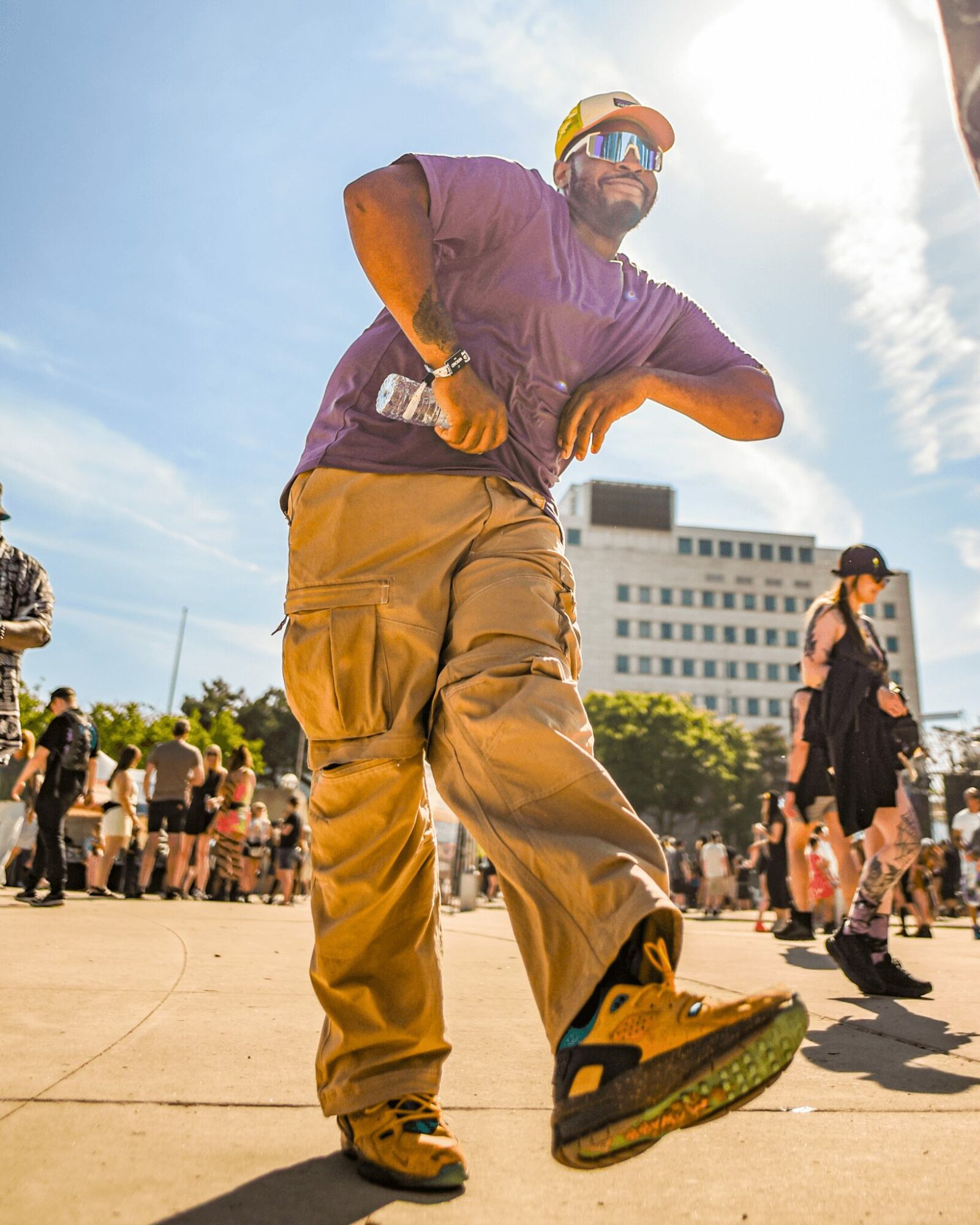 Man in purple shirt and tan pants dancing outdoors with a crowd and a building in the background.