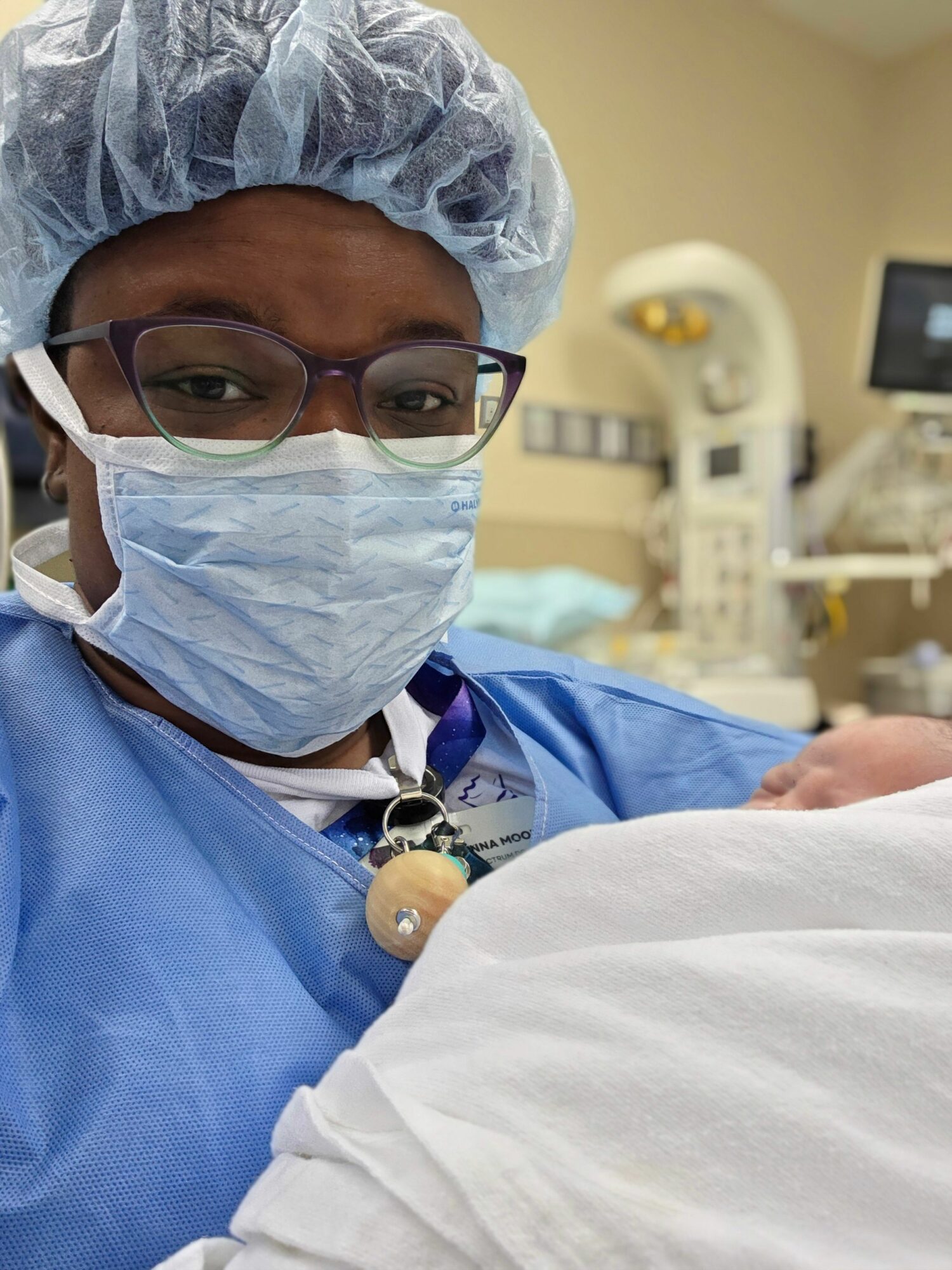Healthcare worker wearing scrubs, mask, glasses, and cap in a clinical setting, holding a patient's hand.