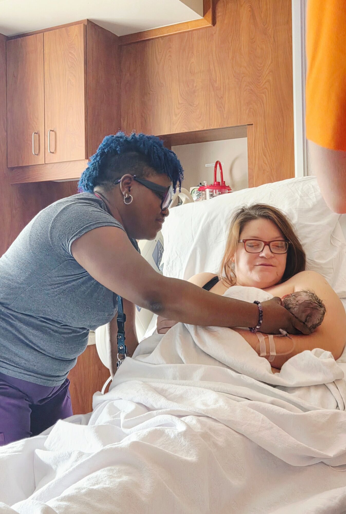 A person with short dark hair and glasses is holding a patient's arm in a hospital room with wooden cabinets.