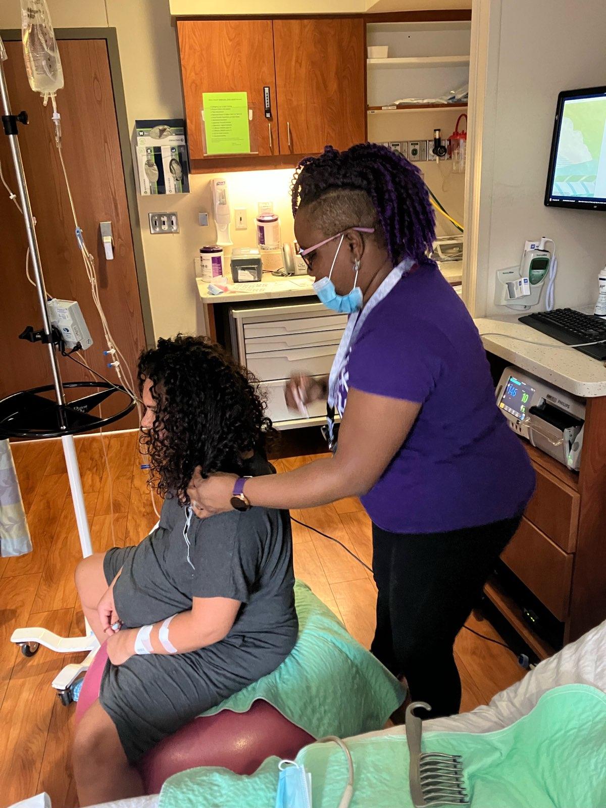 Healthcare worker in purple scrubs and face mask tending to a seated patient with curly hair in a medical room.