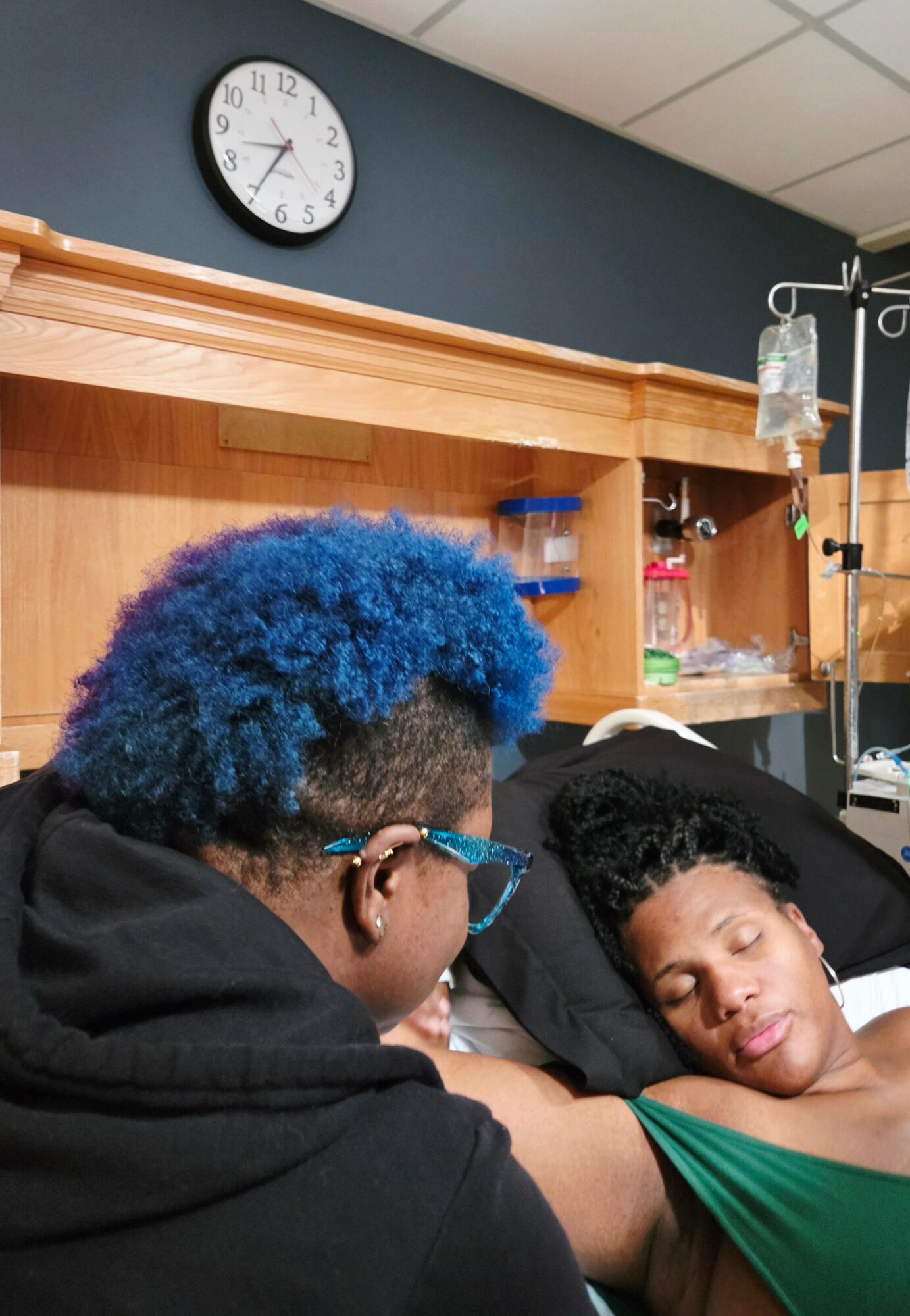Person with blue hair and glasses leaning over a woman lying in a hospital bed, in a medical room.