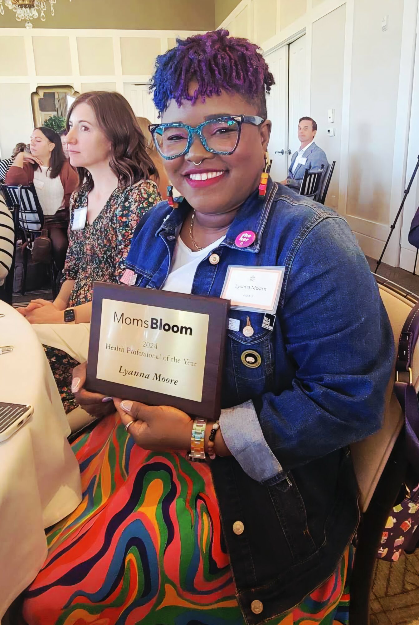Smiling woman with purple hair and glasses holding a framed award at an indoor event, seated at a table.