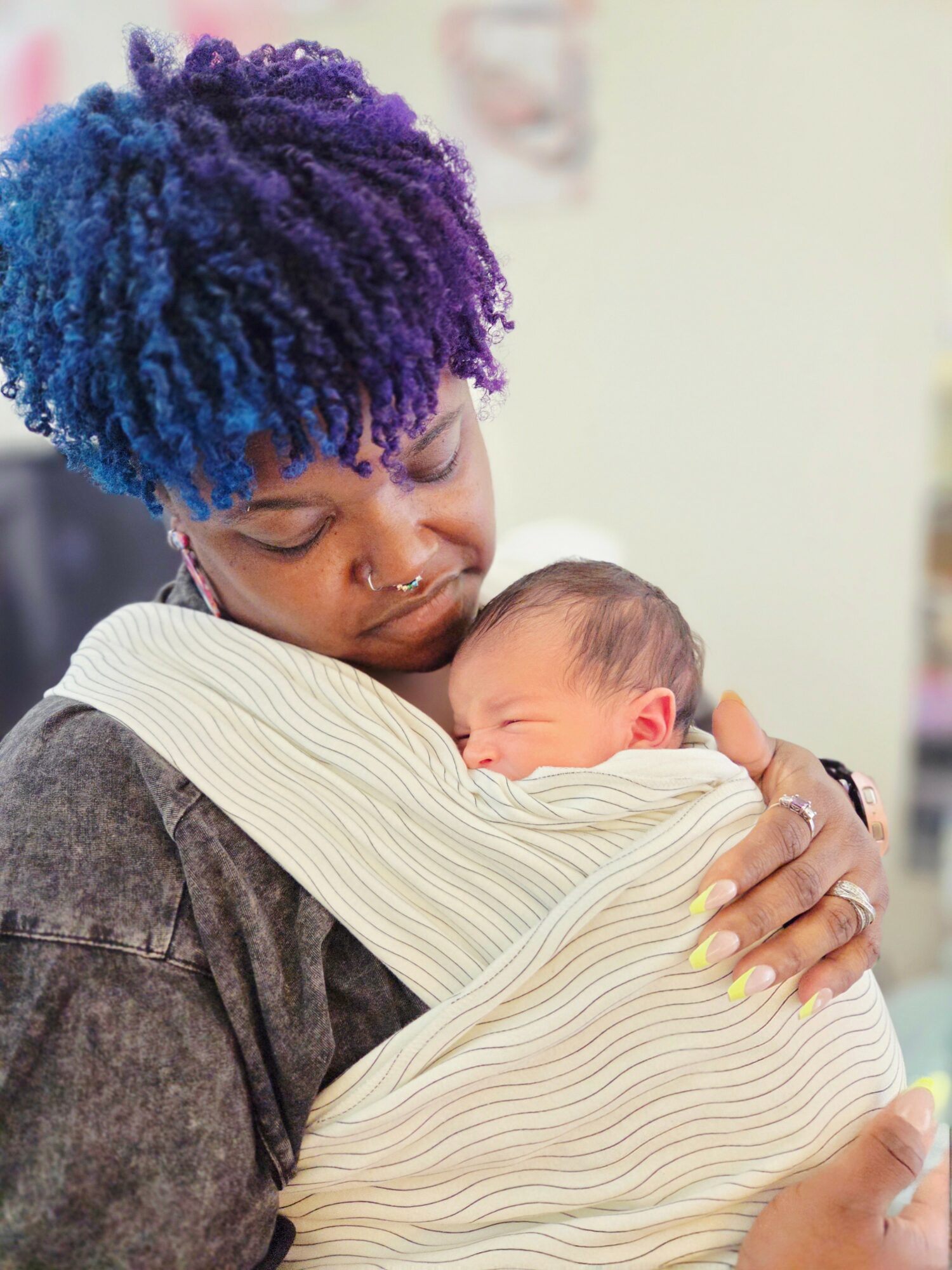 Person with colorful hair holding a sleeping newborn wrapped in a striped blanket.