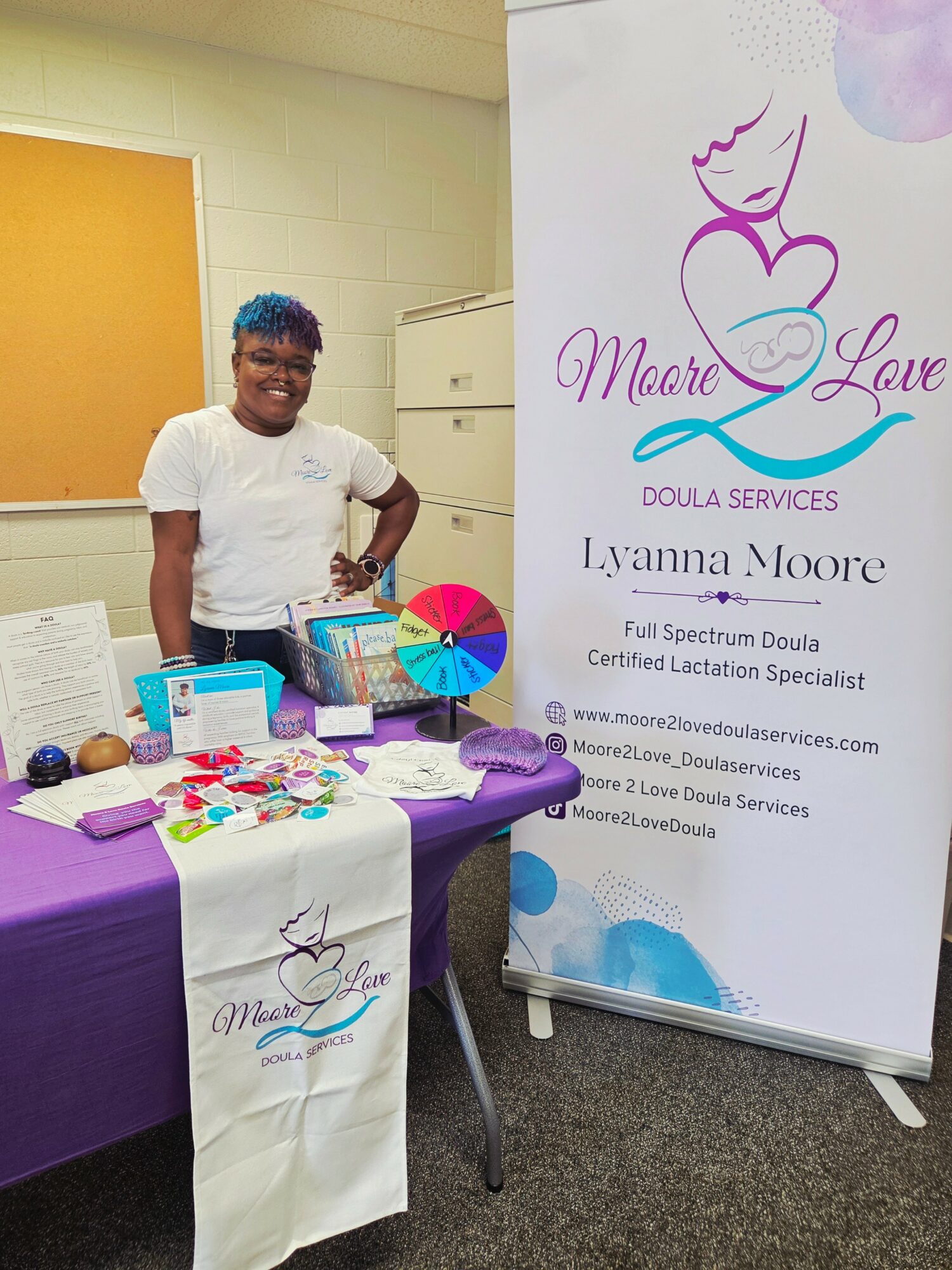 Smiling person with short hair and glasses standing next to a table with colorful items and a banner for Moore Love Doula Services.