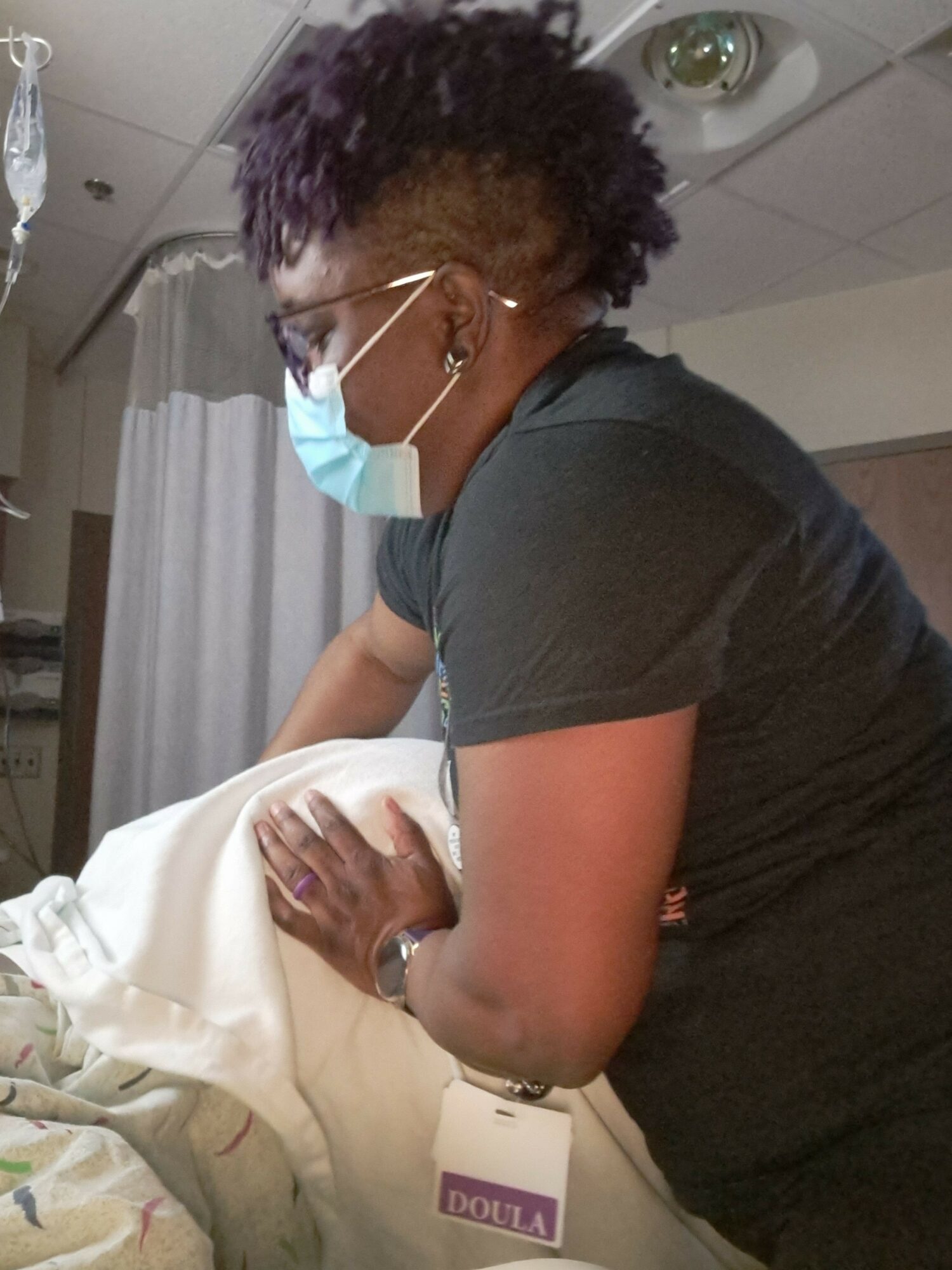 Person wearing a mask and glasses, leaning over a patient in a medical setting.