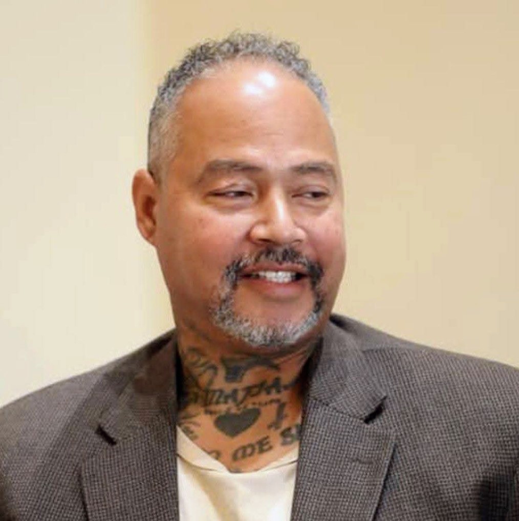 Man with short curly hair, beard, and tattoos on neck, wearing a suit jacket, smiling, against a plain background.