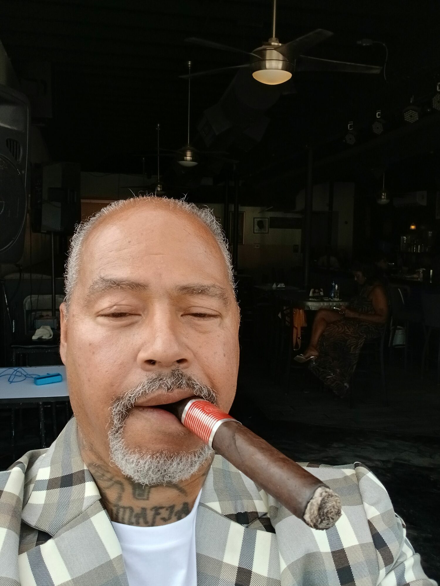 Man with gray beard smokes a cigar, eyes closed, wearing a checkered shirt, indoors with dark background.