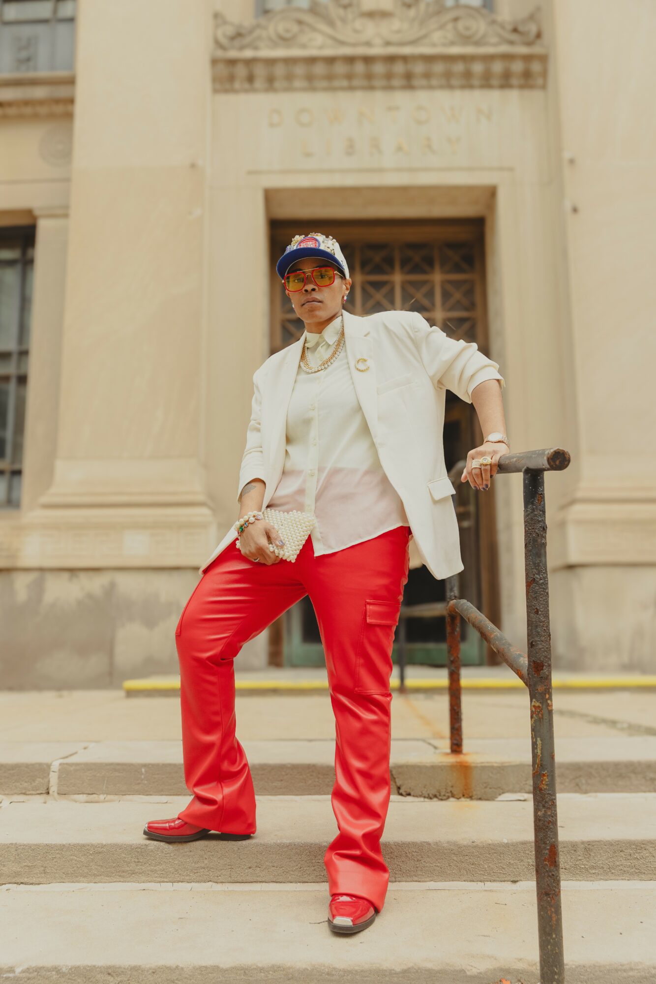 Person standing on steps in front of a building with columns, wearing a white jacket, red pants, and a cap.