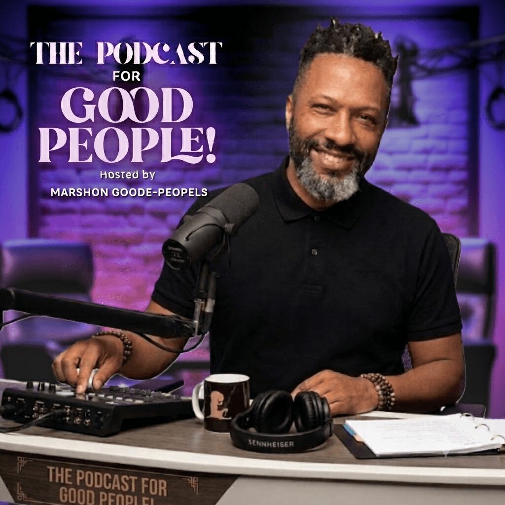 Man with a beard smiling, sitting at a table with podcast equipment, microphone, headphones, and a mug, in a studio setting.