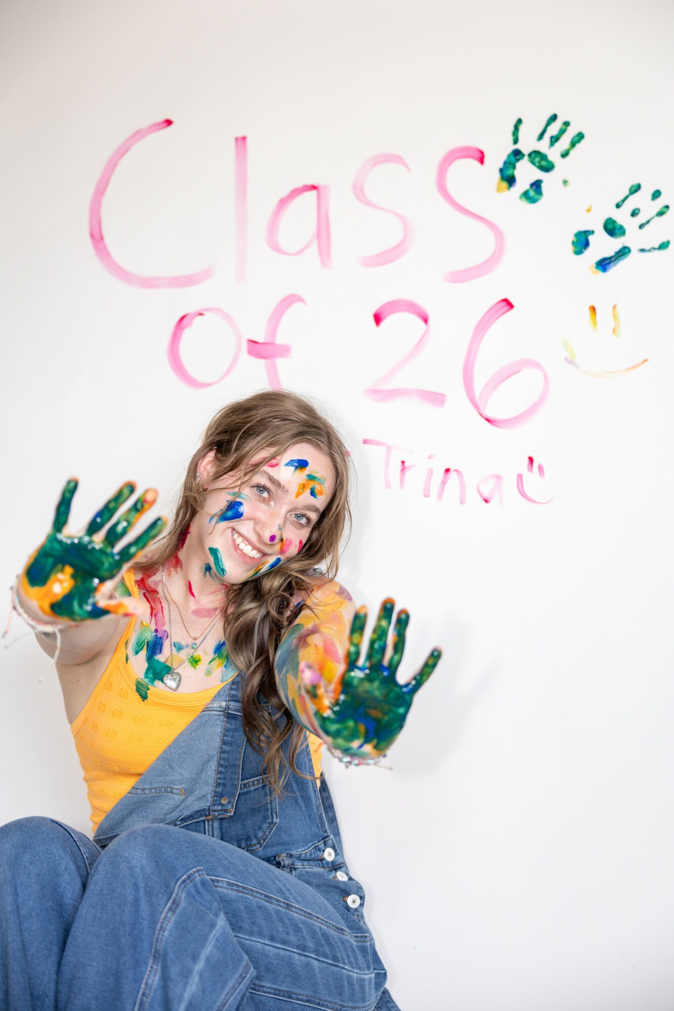 Person with painted hands and face, sitting against a white wall with colorful writing and drawings, smiling and showing hands.