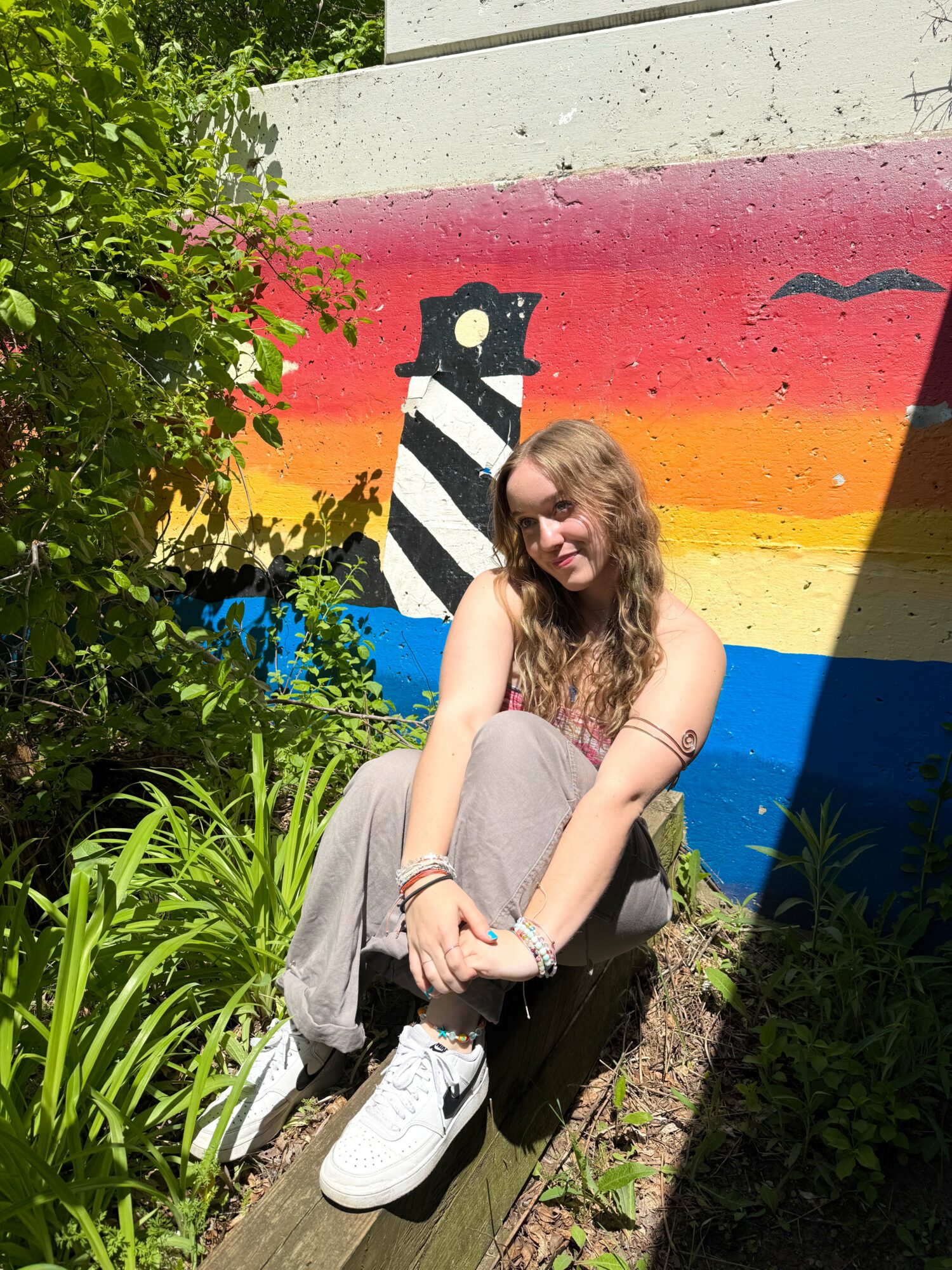 Young woman with long hair sitting on a wooden plank in front of a colorful mural of a lighthouse and sunset, surrounded by greenery.