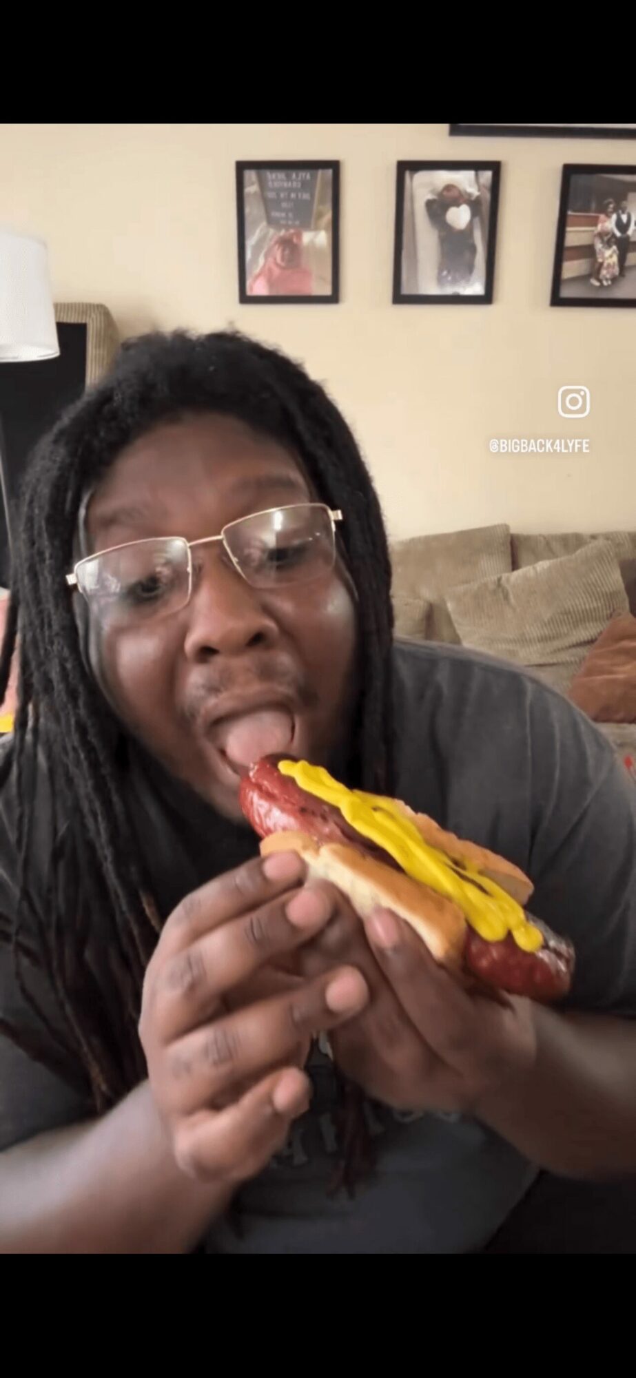 Person with glasses eating a hot dog with yellow mustard, sitting on a couch in a living room, framed pictures on wall