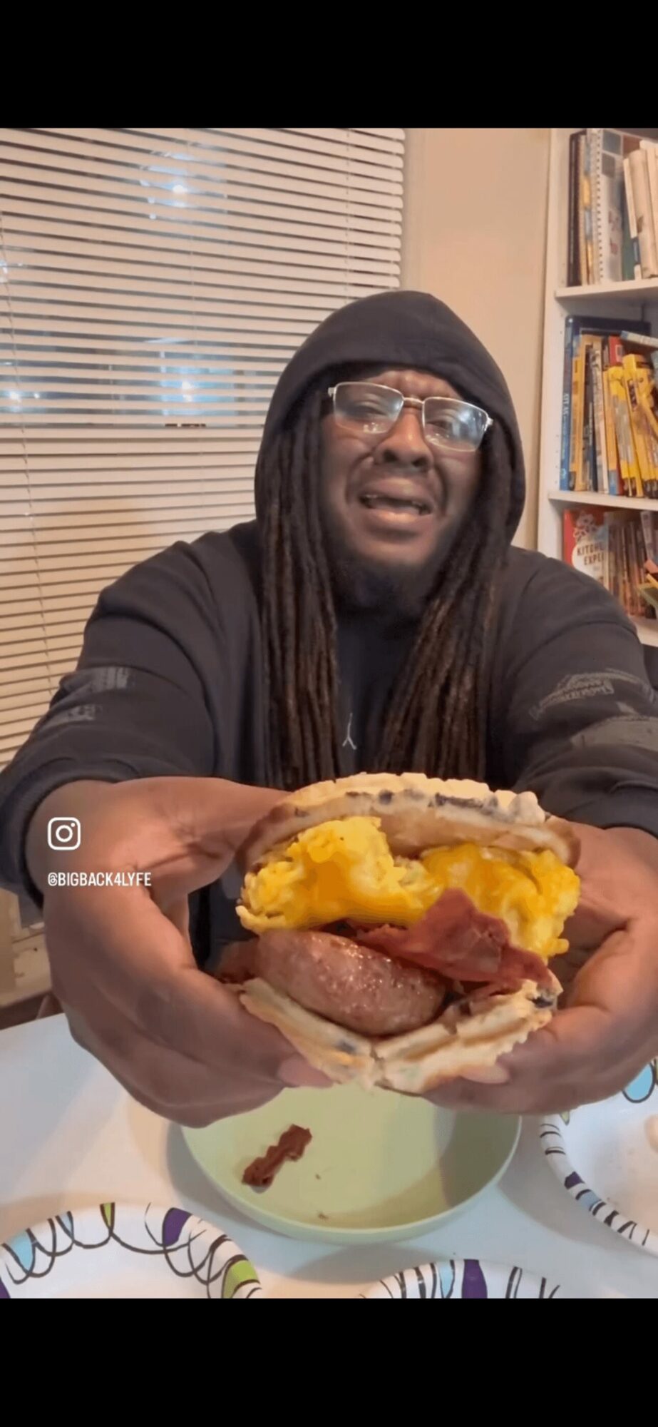 Person with glasses and long hair holds a sandwich with cheese, meat, and vegetables indoors, bookshelf in background.