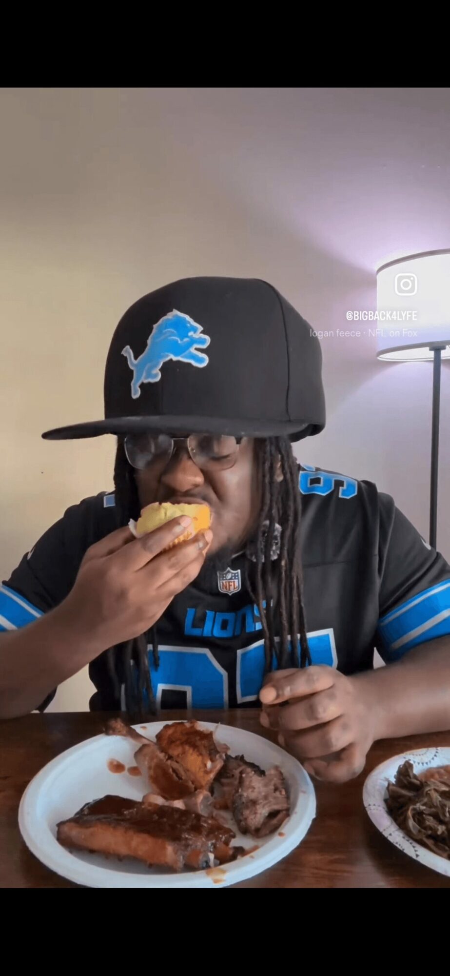 Person wearing a black cap with a blue logo, eating food at a table, with a lamp in the background.
