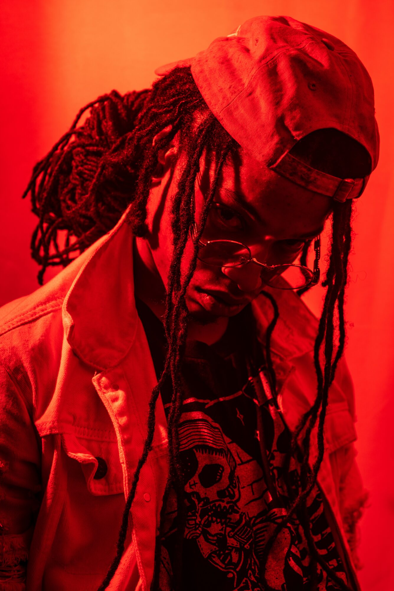 Person with long dreadlocks wearing a red cap, glasses, and a jacket, looking down against a red background.