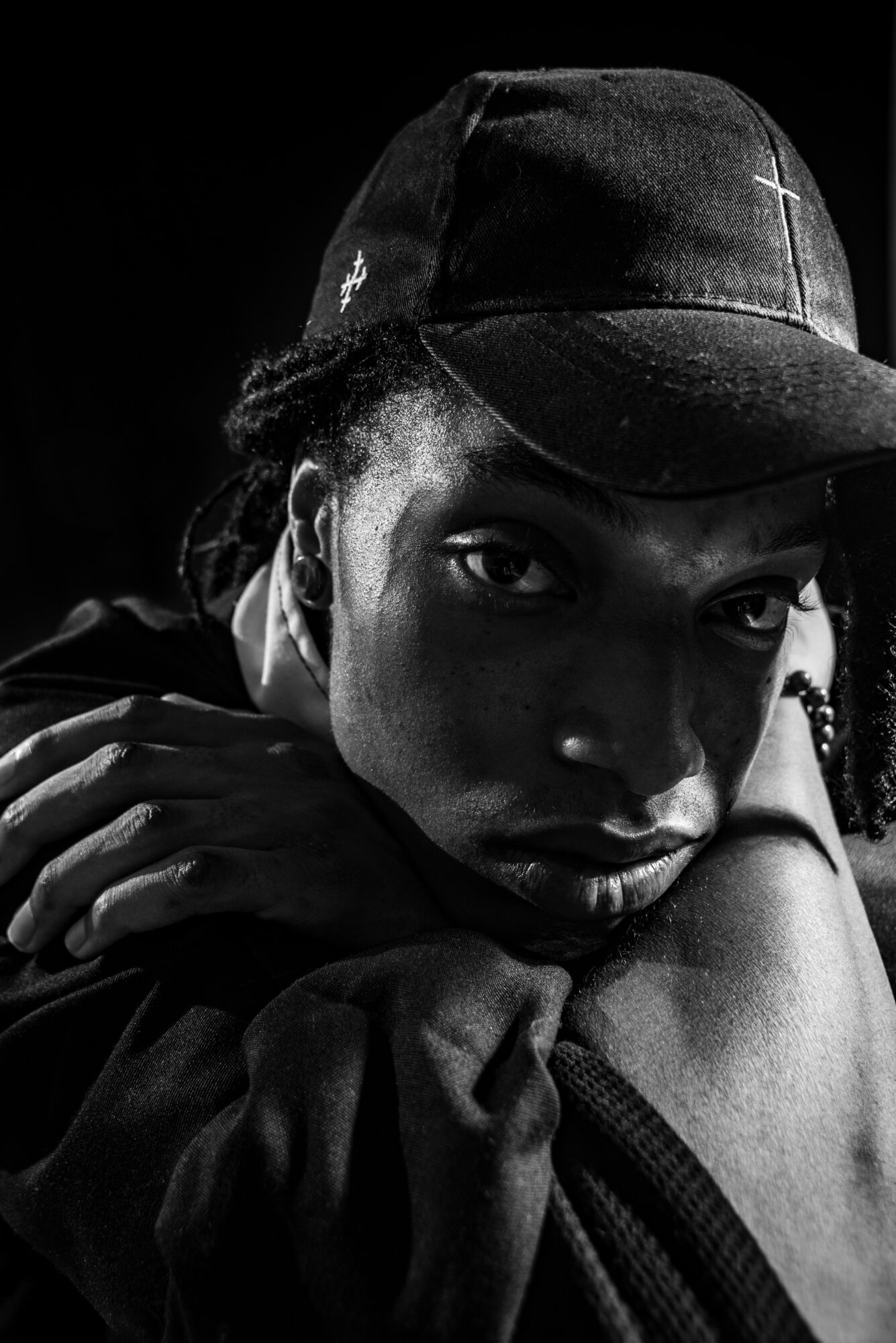 Close-up of a person wearing a cap, resting their head on their arm, with a serious expression, in black and white.