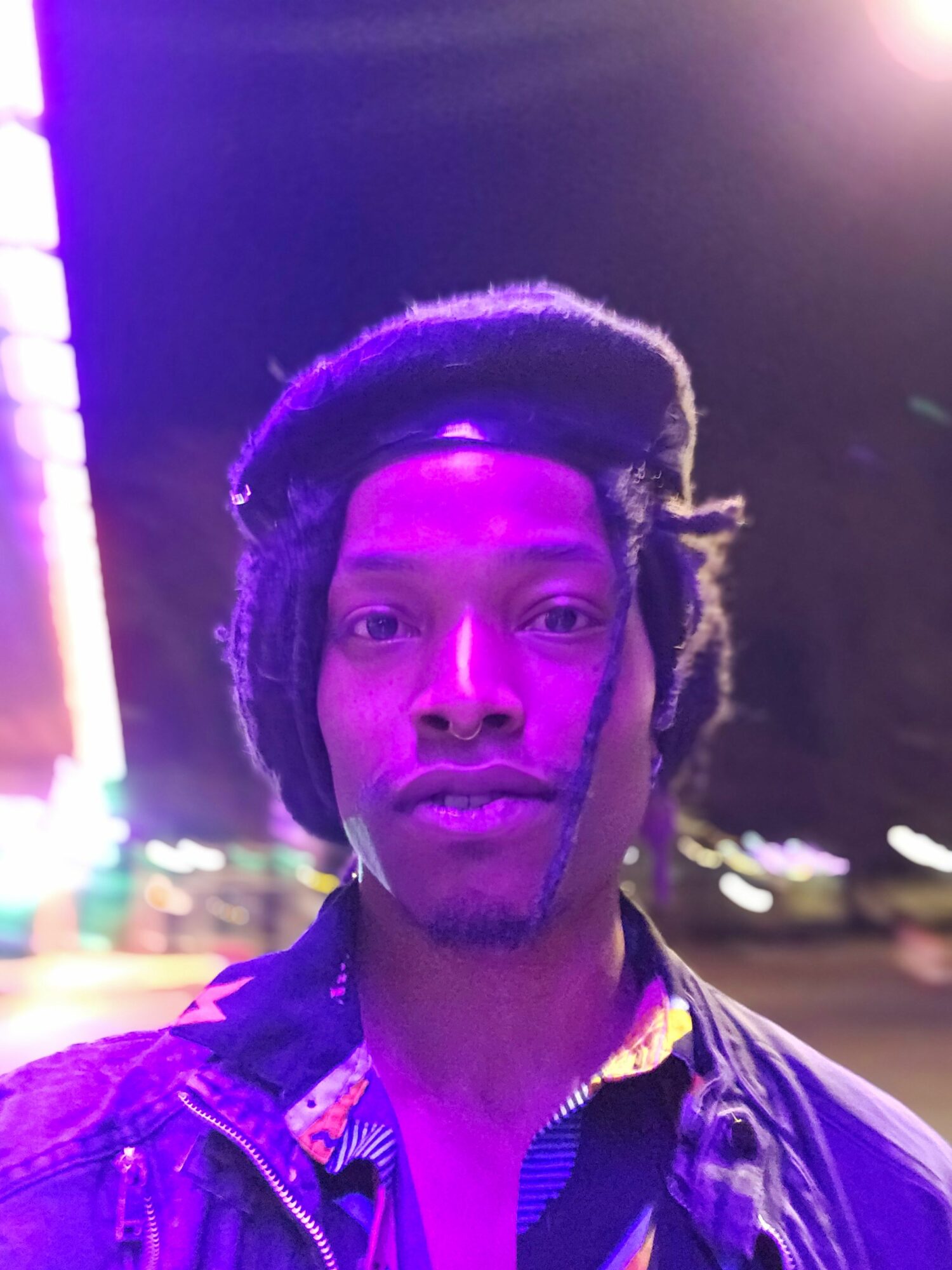 Young man with dreadlocks and a serious expression, illuminated by colorful neon lights, facing forward in a nighttime setting.