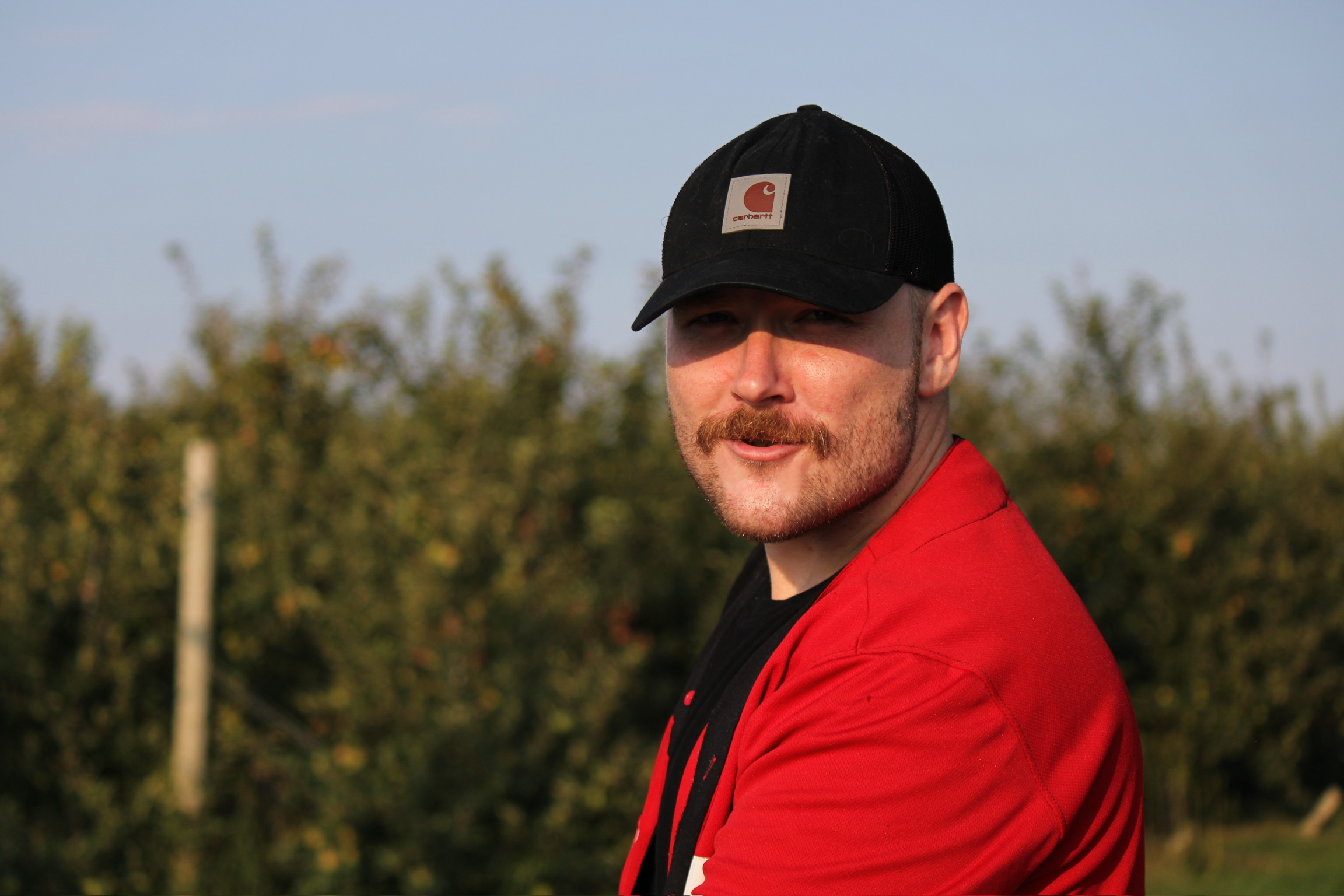 Man wearing a black cap and red jacket outdoors, with trees and sky in the background.