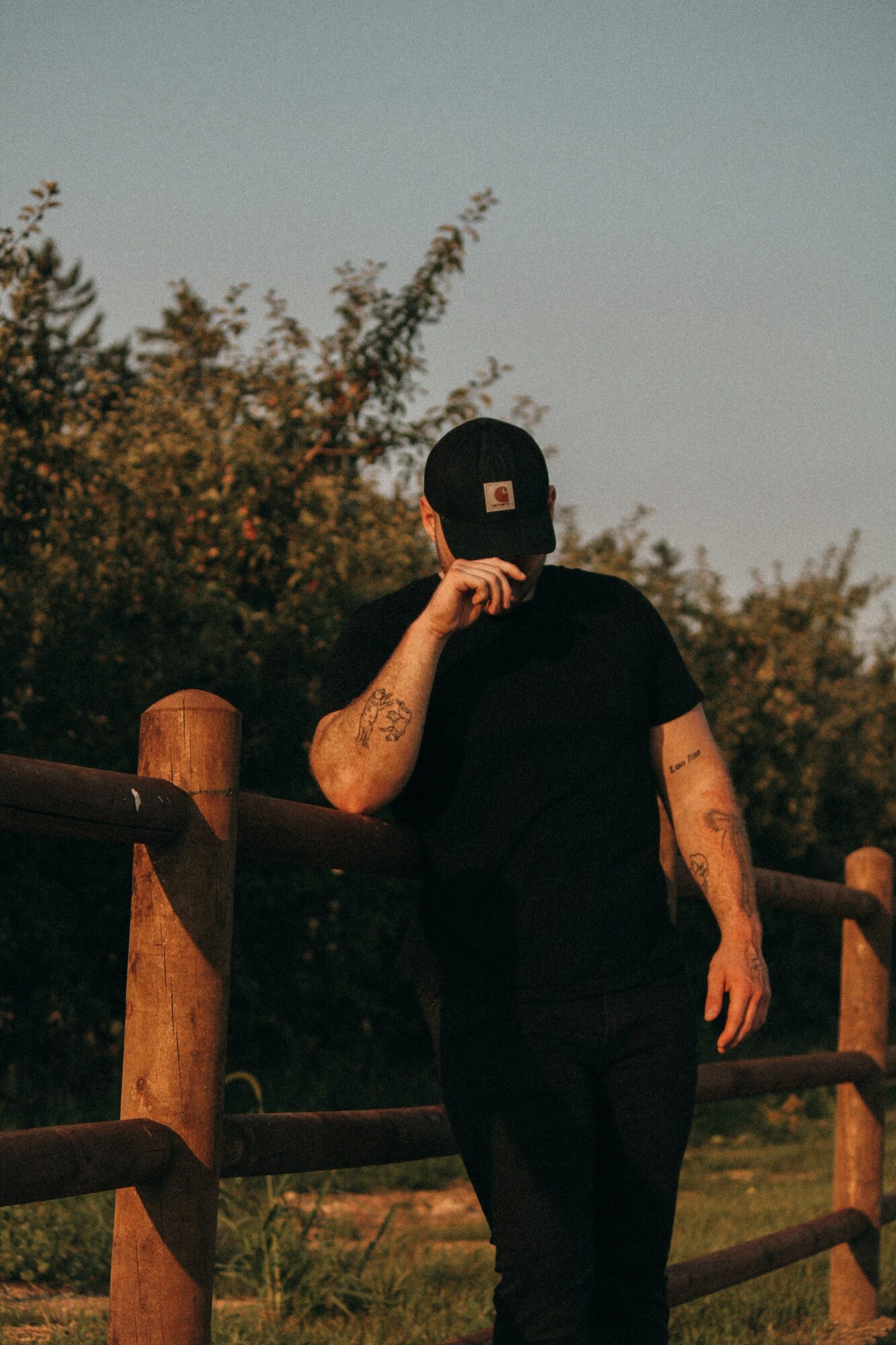 Person wearing a black cap and shirt, standing outdoors near a wooden fence, with trees and sky in the background.