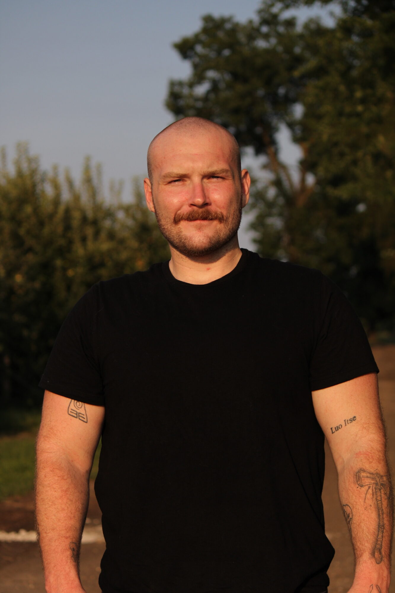 Man with a beard and tattoos on arms, wearing a black shirt, outdoors with trees in background.