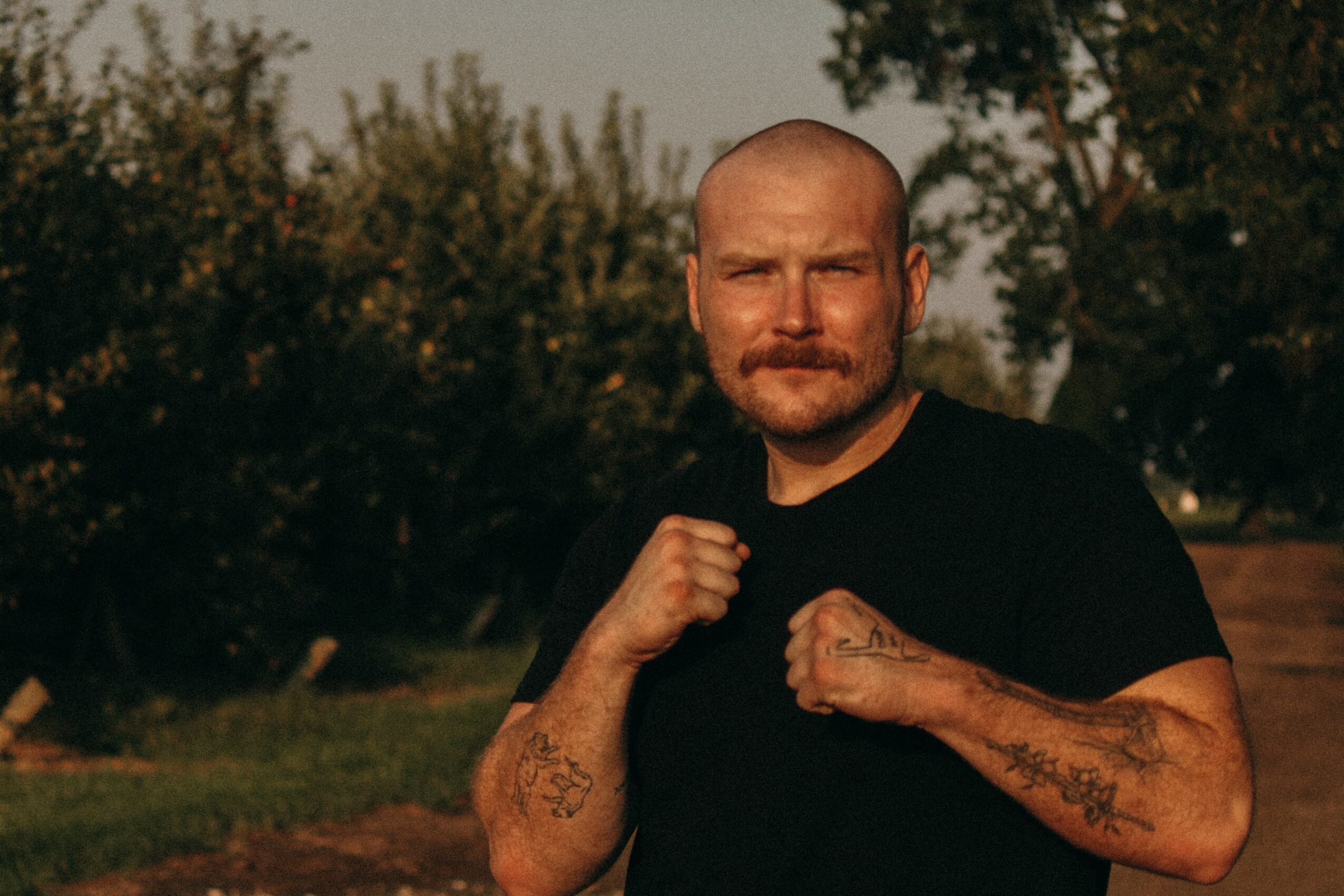 Man with a beard and tattoos on arms, wearing a black shirt, outdoors with trees in background.