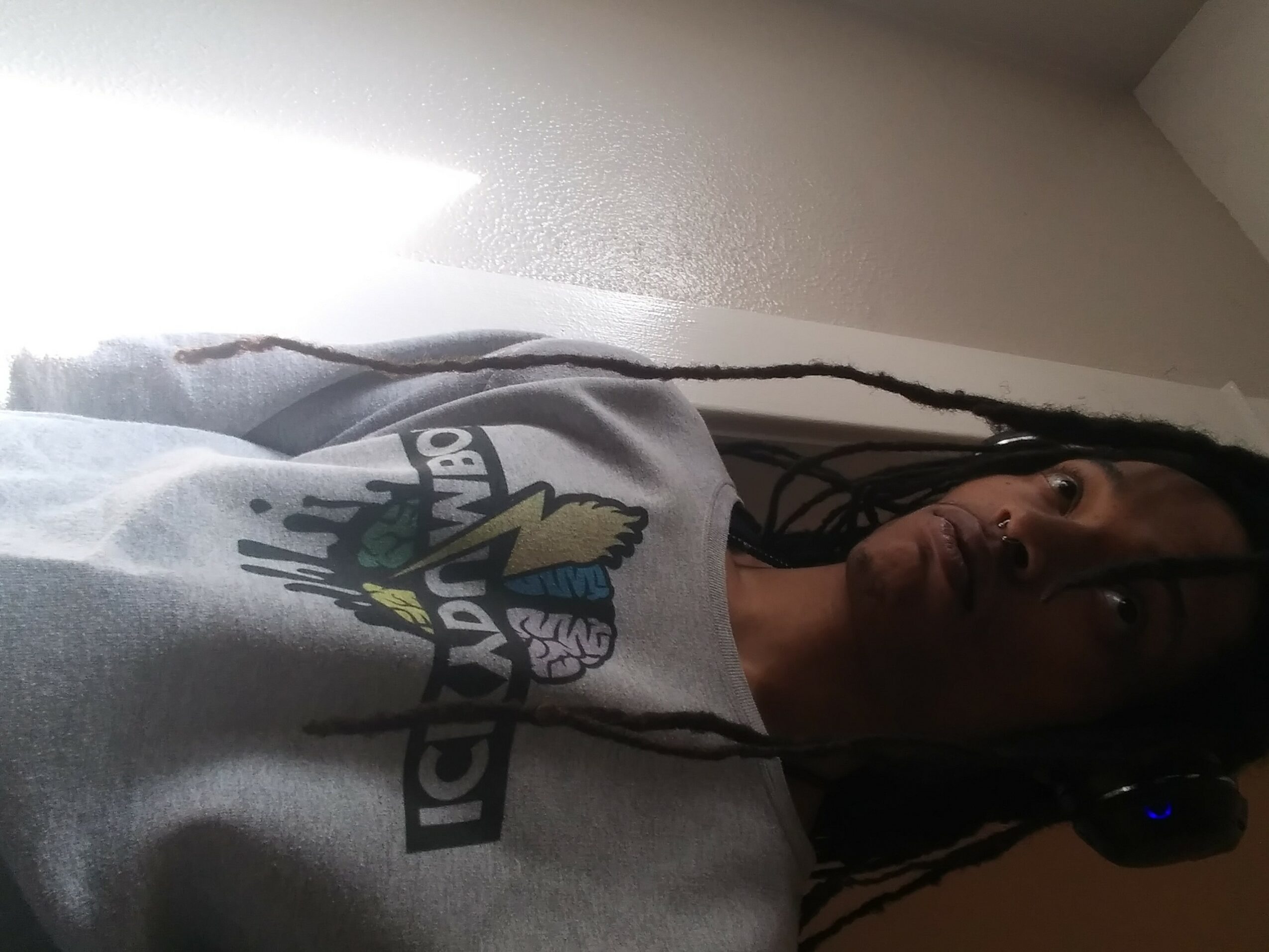 Person with long braided hair wearing a gray T-shirt with a graphic design, indoors with light coming from the left.