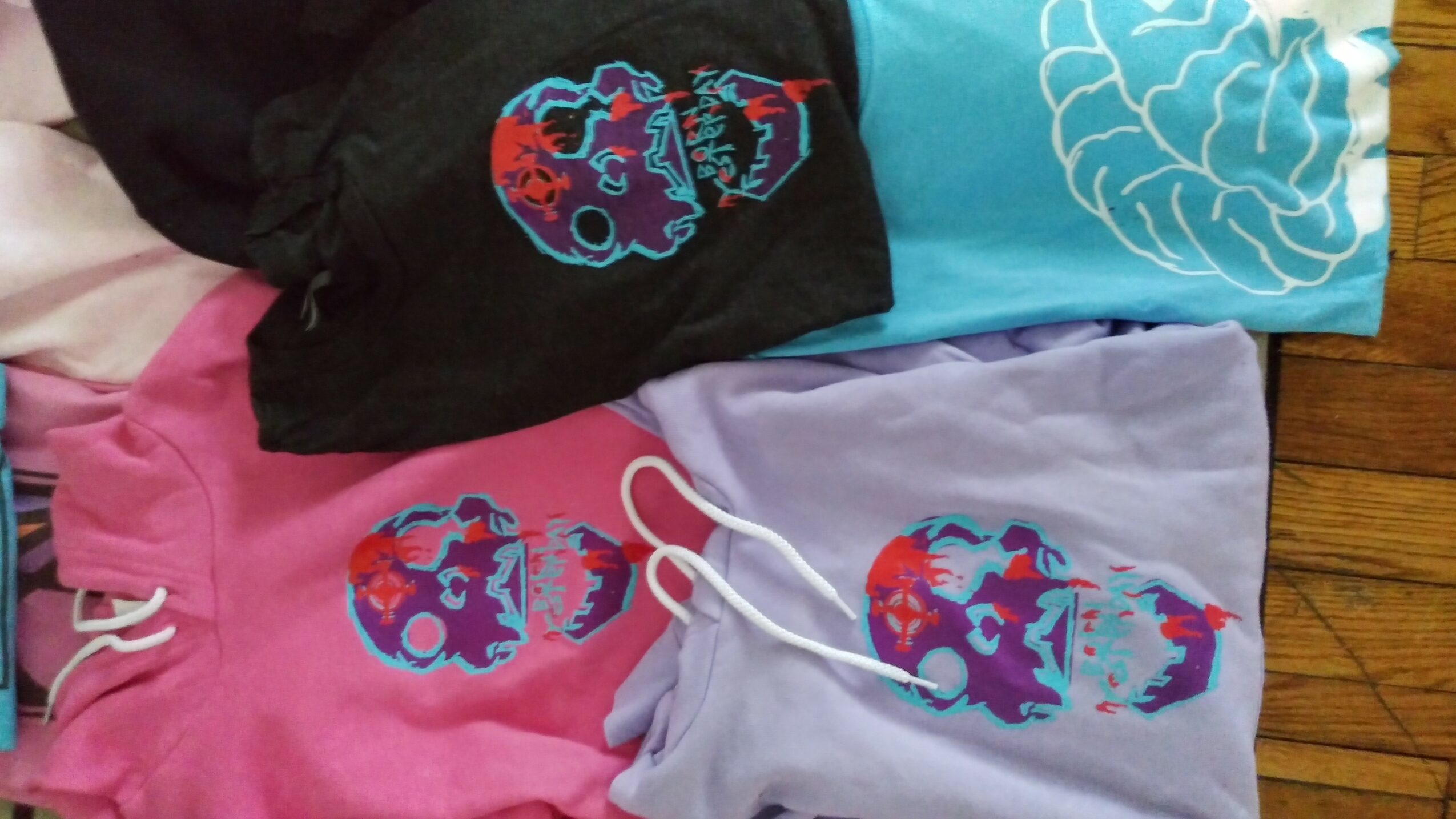 Four folded T-shirts with colorful skull designs, in black, pink, lavender, and light blue, on a wooden surface.