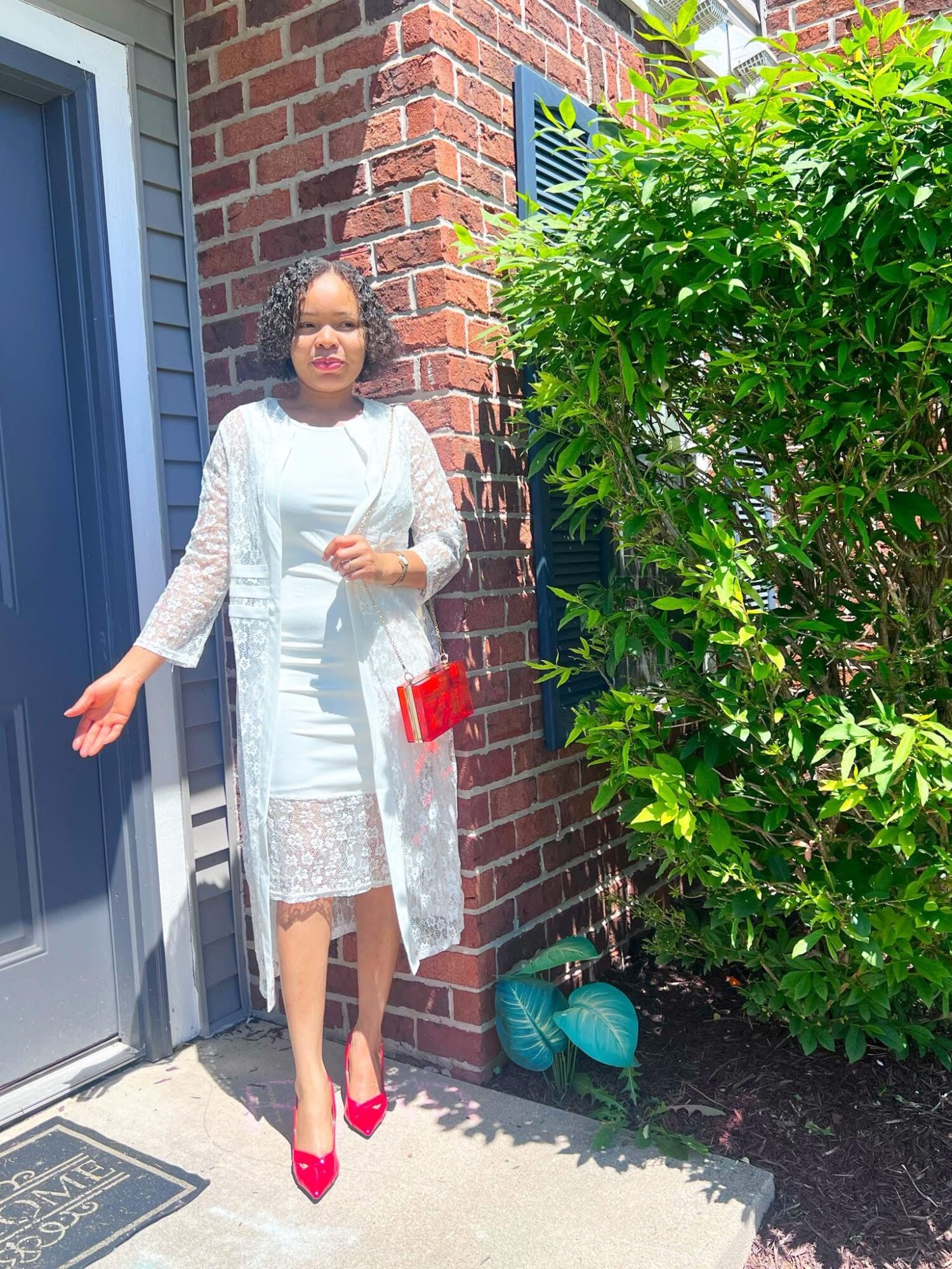 Woman in white dress and lace coat standing outside near a brick wall and green bush, sunlight shining.