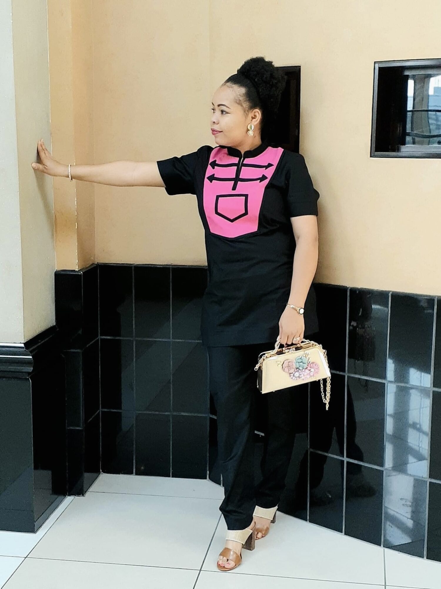 Woman in black outfit with pink design, standing against wall, holding a purse, extending her arm, in an indoor setting.