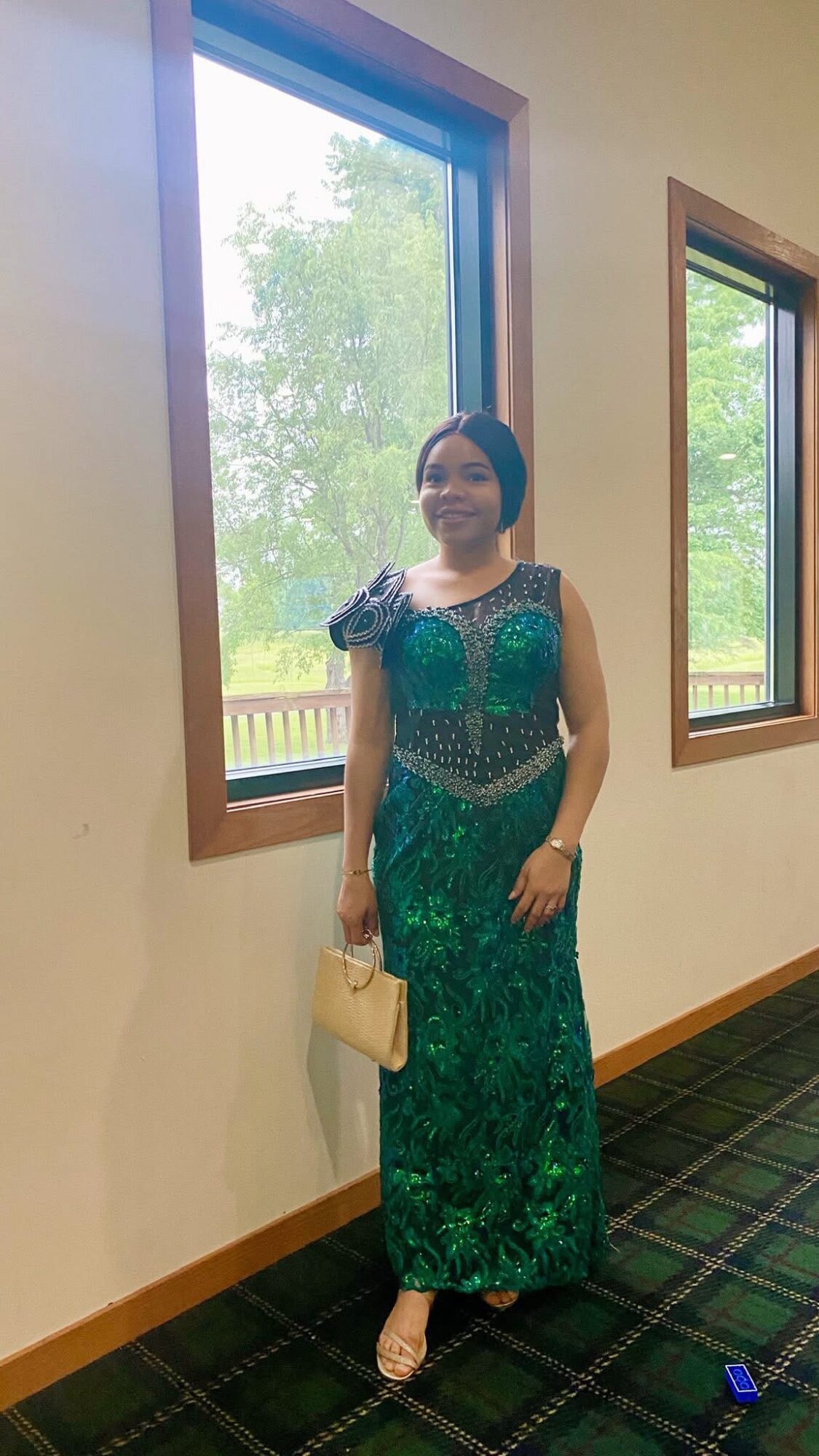 Woman in a green dress standing indoors near windows with trees outside, holding a beige purse, smiling.
