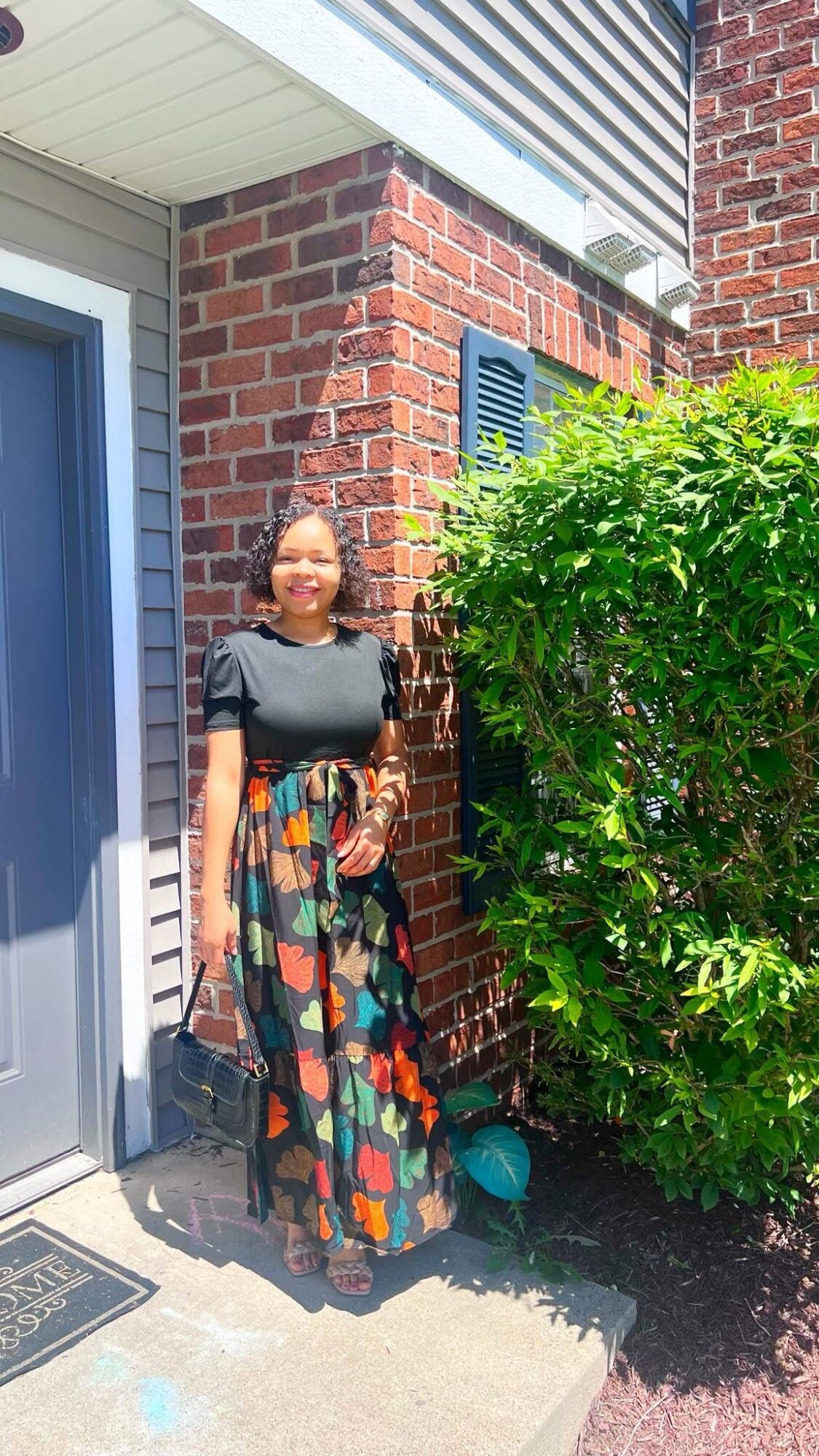 Woman standing outside next to a brick wall and green bush, wearing a black top and colorful long skirt, smiling.
