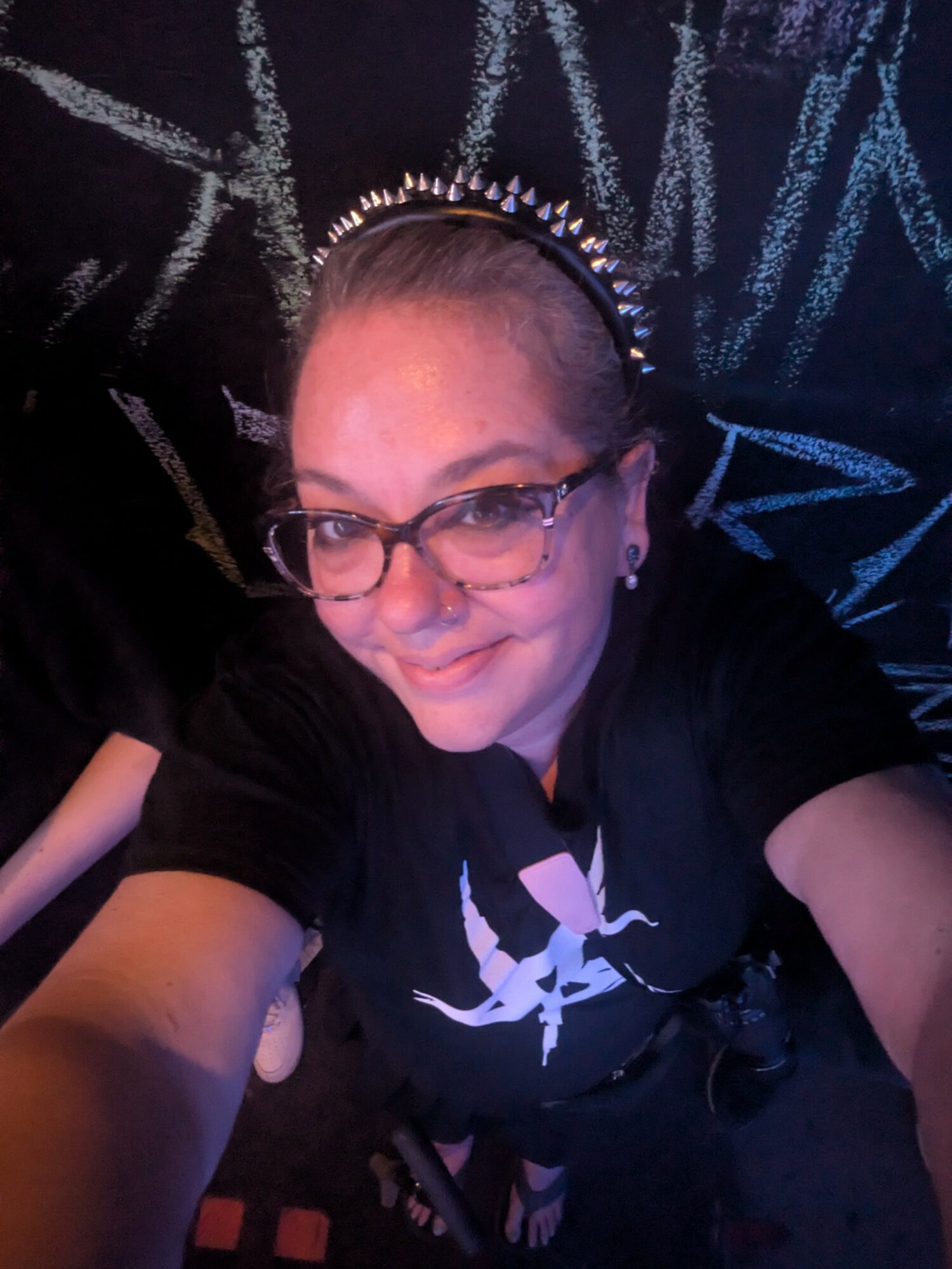 Person with glasses and a headband, smiling, taking a selfie in front of a chalkboard with writing.