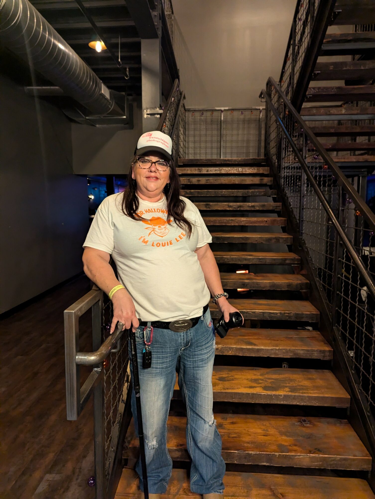 Woman standing on a staircase holding a camera, wearing a white t-shirt, jeans, and a cap, indoors with industrial decor.