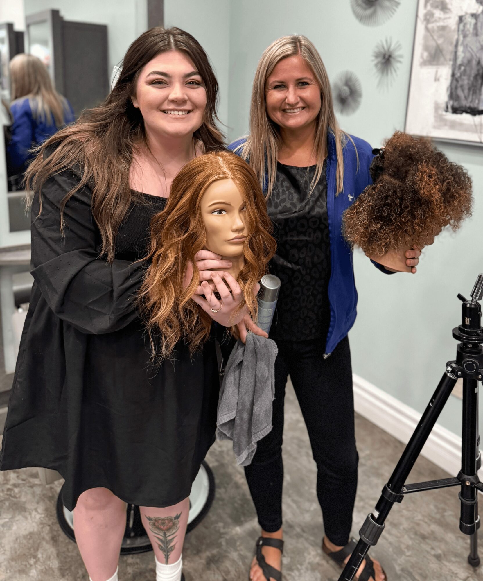 Two women smiling, one holding a mannequin head with red hair, in a salon or studio setting, with hair and art on walls.