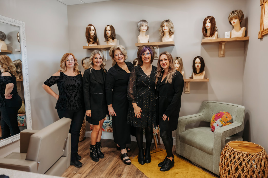 Five women stand together in a room with mannequin heads on shelves behind them, some smiling and posing for the photo.