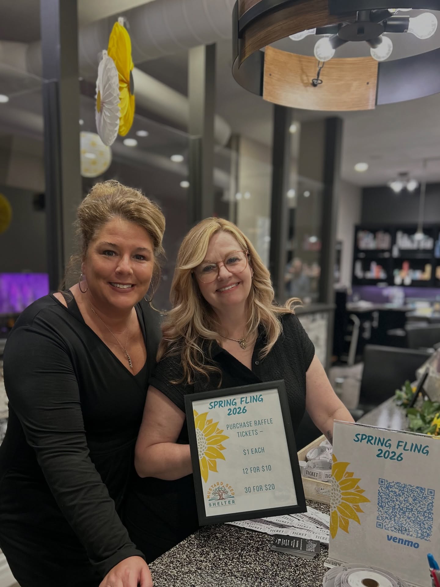 Two women smiling behind a table with a framed certificate and a sign, in a modern indoor setting.