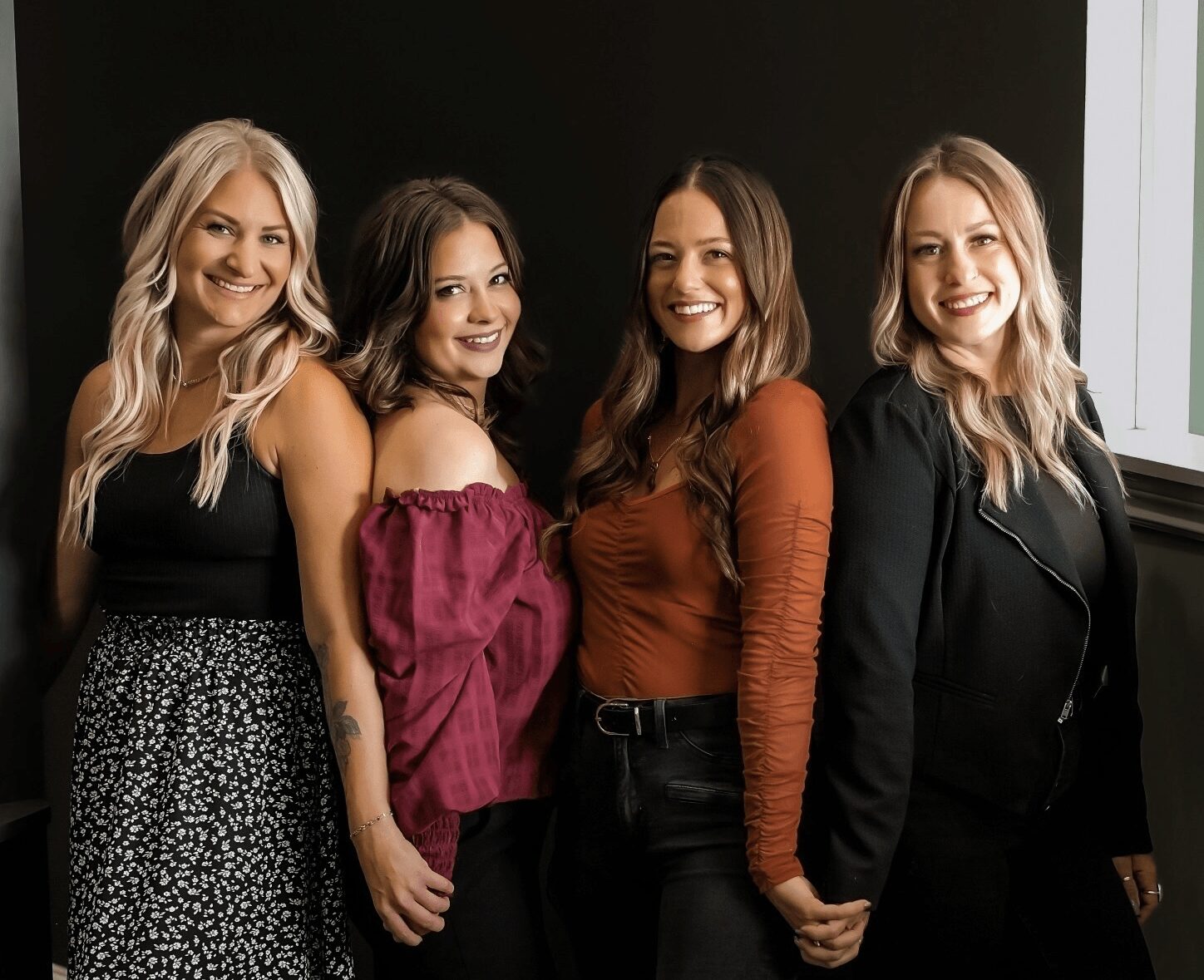 Four women standing side by side, smiling, in front of a dark background with a window on the right.