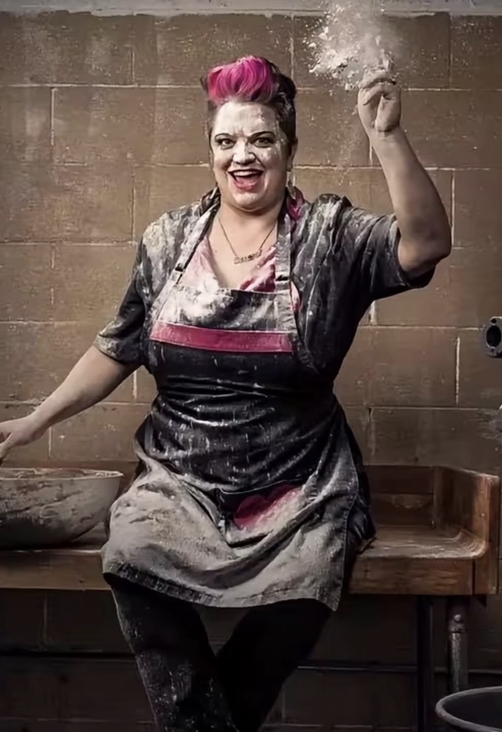 Person with pink hair smiling, throwing flour in the air, wearing an apron, standing in front of a brick wall.