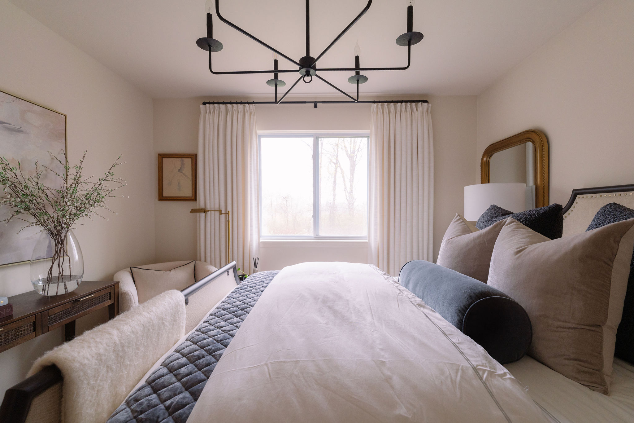 Bedroom with bed, window, curtains, and decorative pillows, illuminated by natural light, with a chandelier overhead.