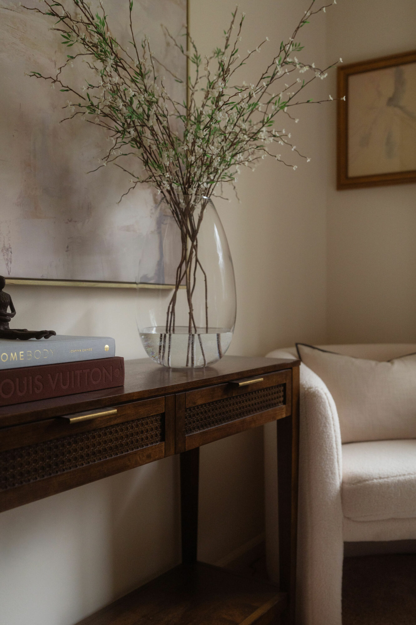 Vase with tall, thin branches and small leaves on a wooden table, with a sofa and framed artwork in the background.