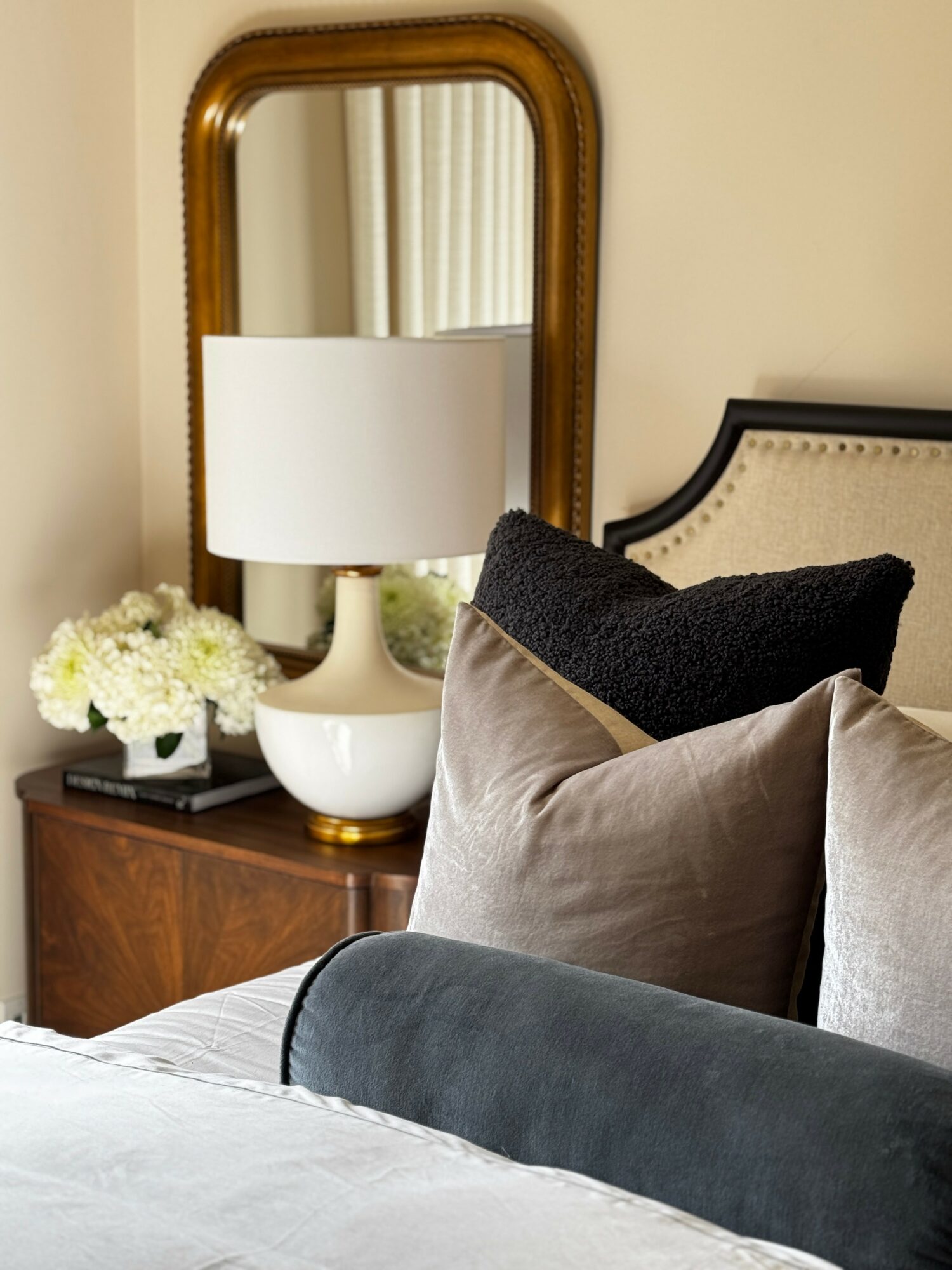 Bedroom corner with a wooden nightstand, white lamp, mirror, and pillows on the bed, including black, beige, and gray pillows.